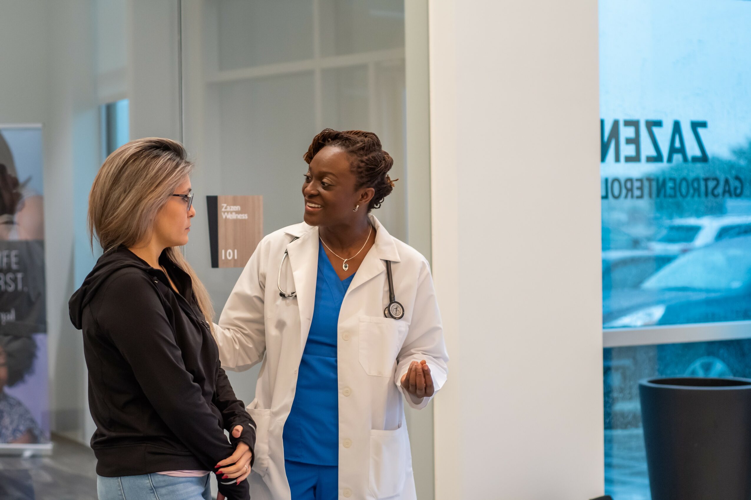 Two women, one in a white coat and blue scrubs, talking in a medical setting, with a window and a sign in the background.