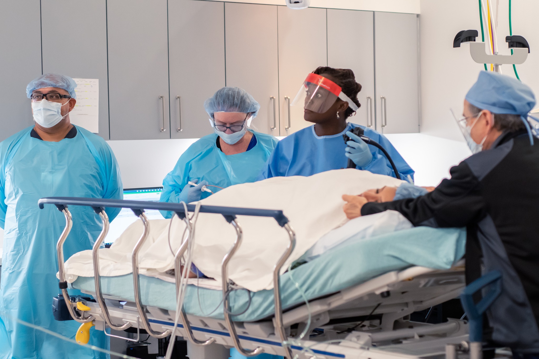 Medical team in scrubs and masks around a hospital bed with a patient, in a clinical setting.