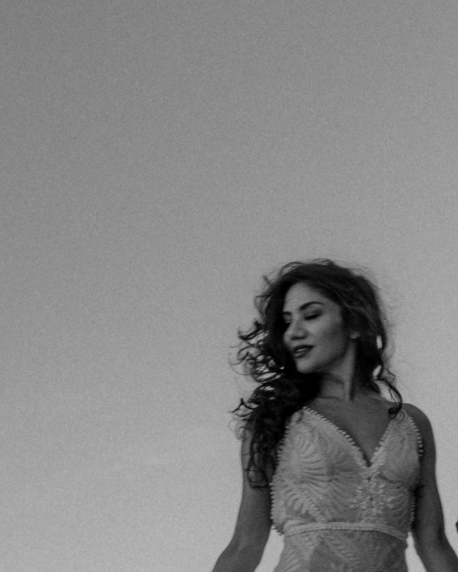 A woman with curly hair stands outdoors against a cloudy sky, looking to the side with a slight smile.