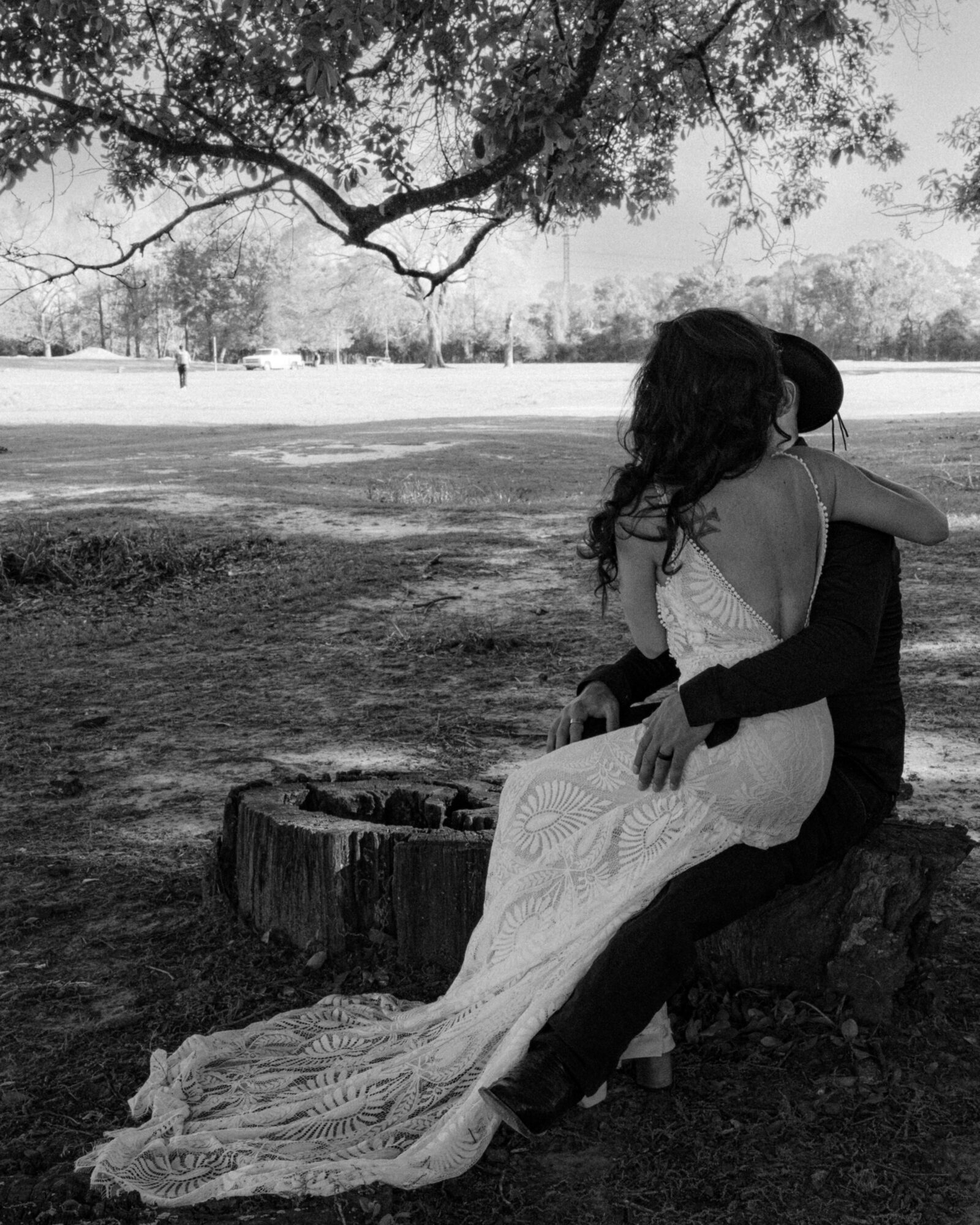 Two people sit on a tree stump embracing outdoors, with a large tree and open field in the background.