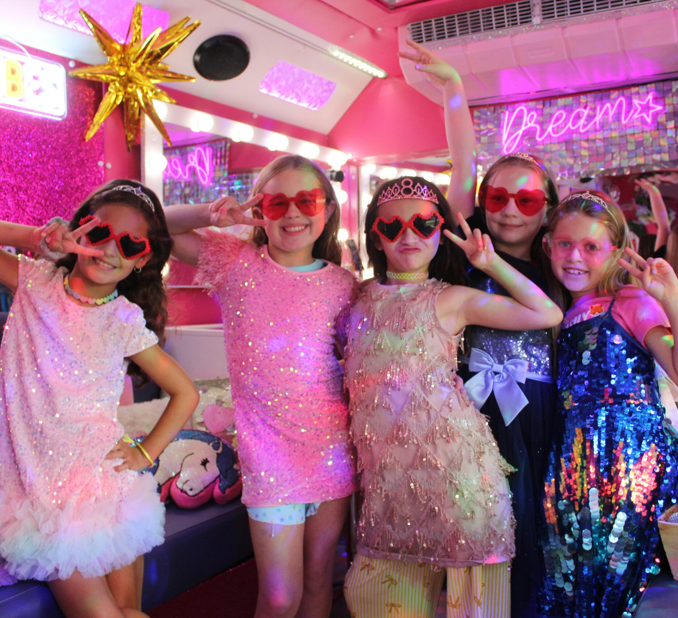 Five young girls in colorful dresses and sunglasses pose in a decorated party room with pink and neon signs.