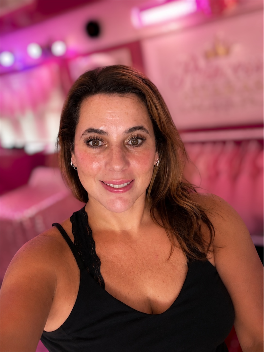 Woman with shoulder-length brown hair smiling in a pink-lit room, wearing a black top with lace straps.