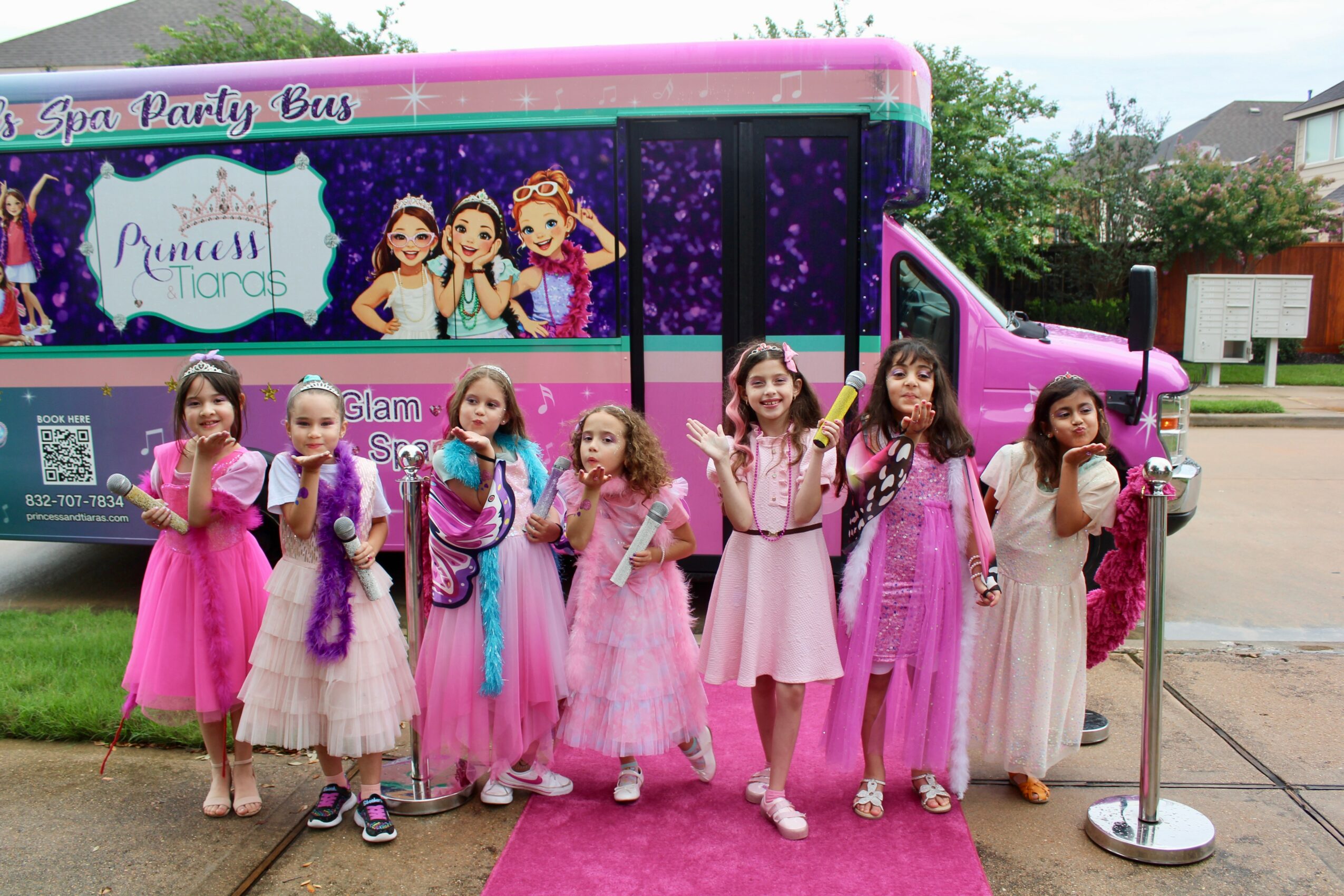 Group of young girls in pink dresses standing in front of a princess-themed party bus, smiling and holding party favors.