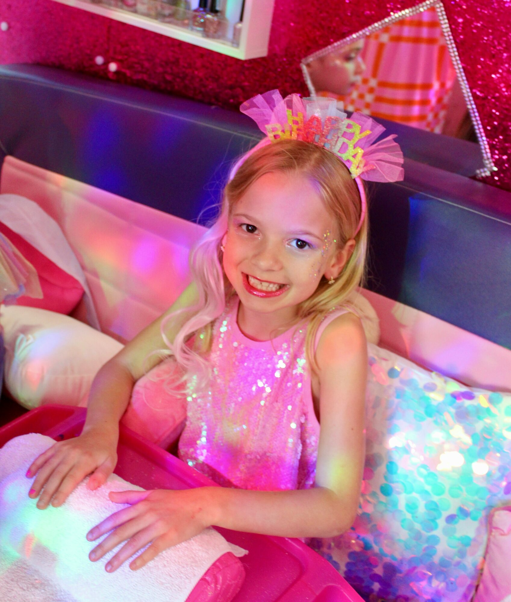 Young girl with a birthday crown, smiling, sitting on a bed with colorful pillows, pink dress, birthday decorations in background.