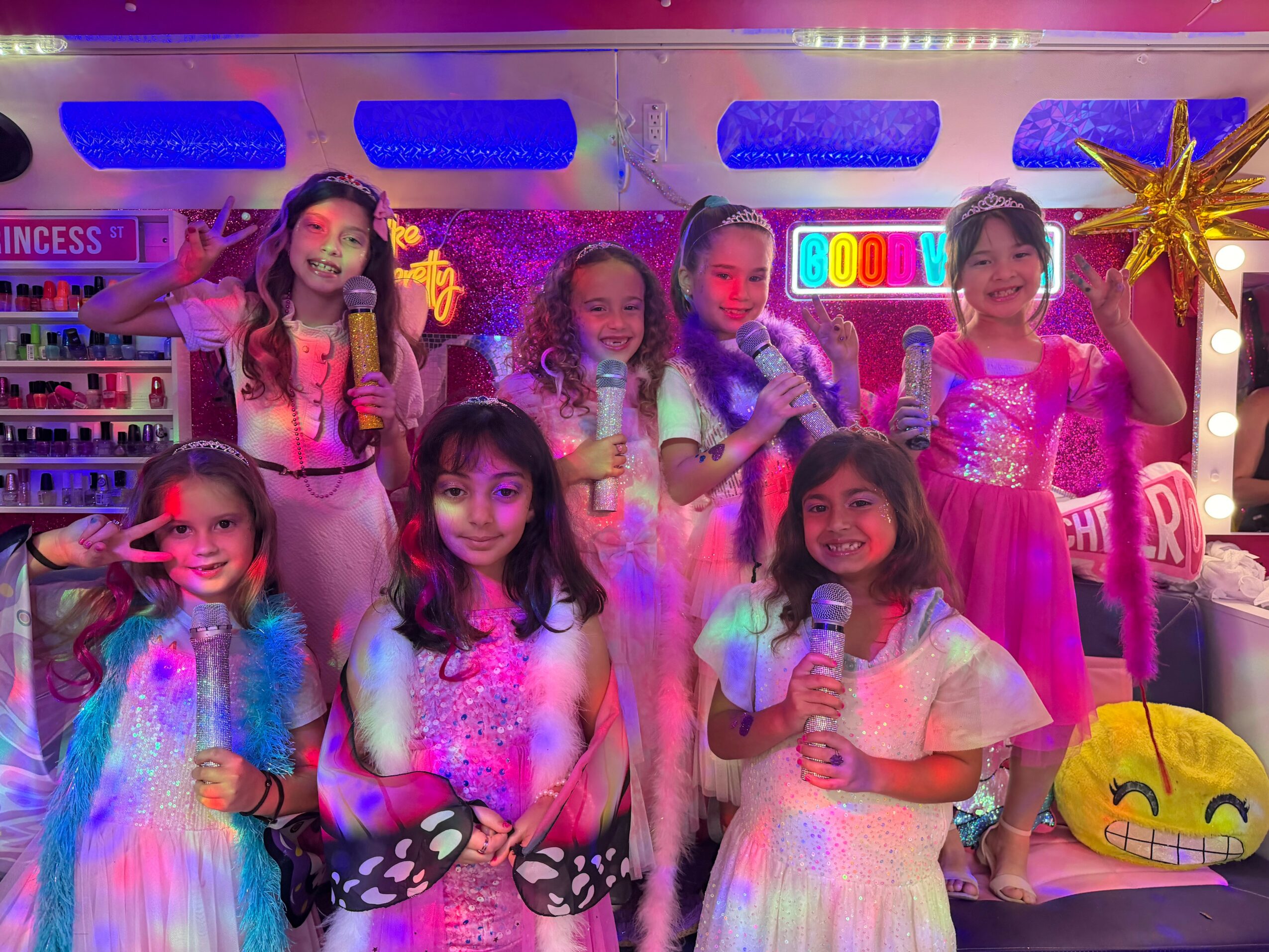 Group of seven children in colorful costumes at a party, some holding microphones, with bright decorations and a smiley face pillow.