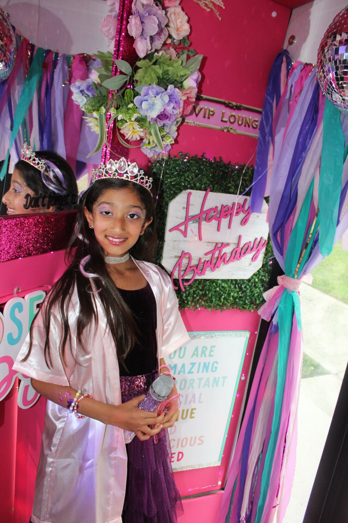 Young girl in a tiara and sash at birthday celebration, standing next to a decorated display with flowers and a birthday sign.