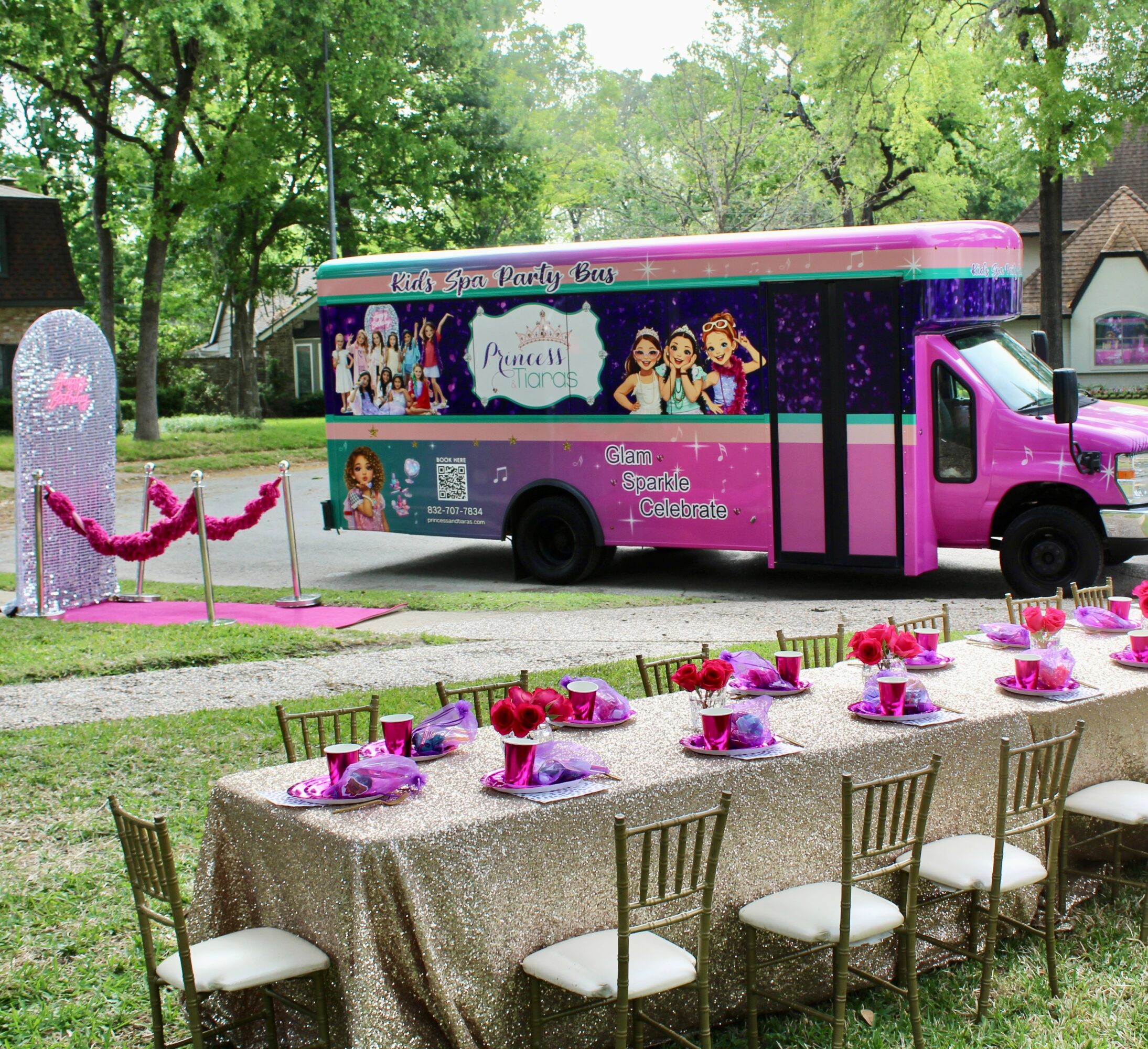 Pink party bus with princess characters parked near decorated table and chairs outdoors, surrounded by trees and greenery.