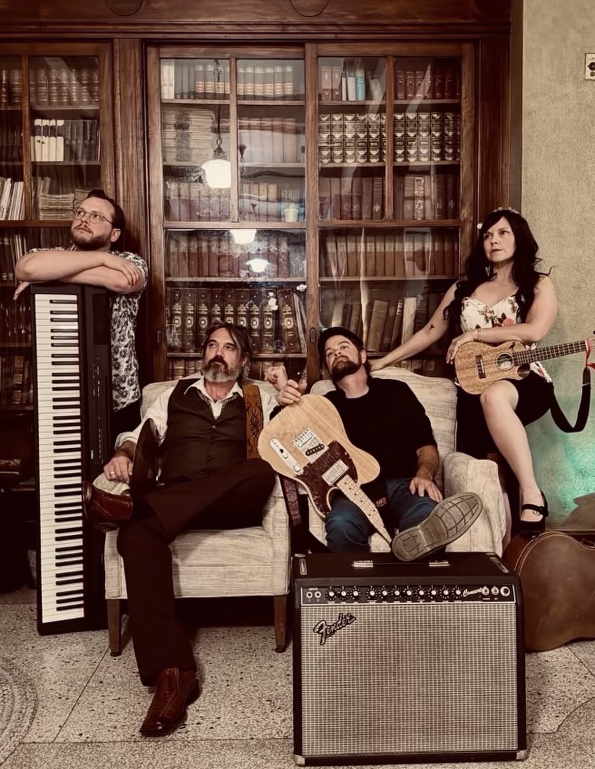 Four people with musical instruments sit in front of a large wooden bookshelf filled with books, in a cozy room.