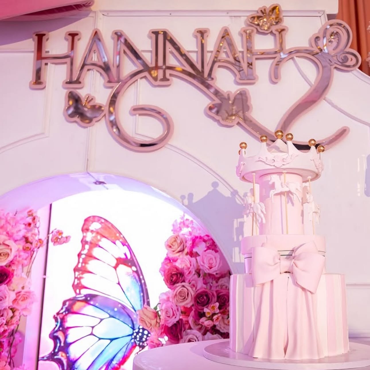 Decorative birthday cake with pink ribbons and a butterfly backdrop, with a sign reading 'Hannah' and butterfly decorations.