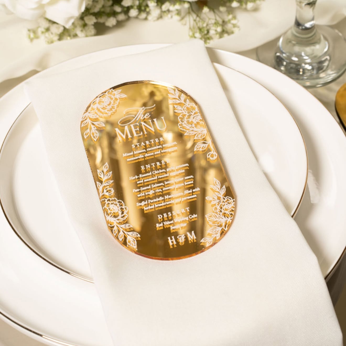 Gold-colored menu card with floral design, placed on a white napkin on a plate, at a formal dining setting.