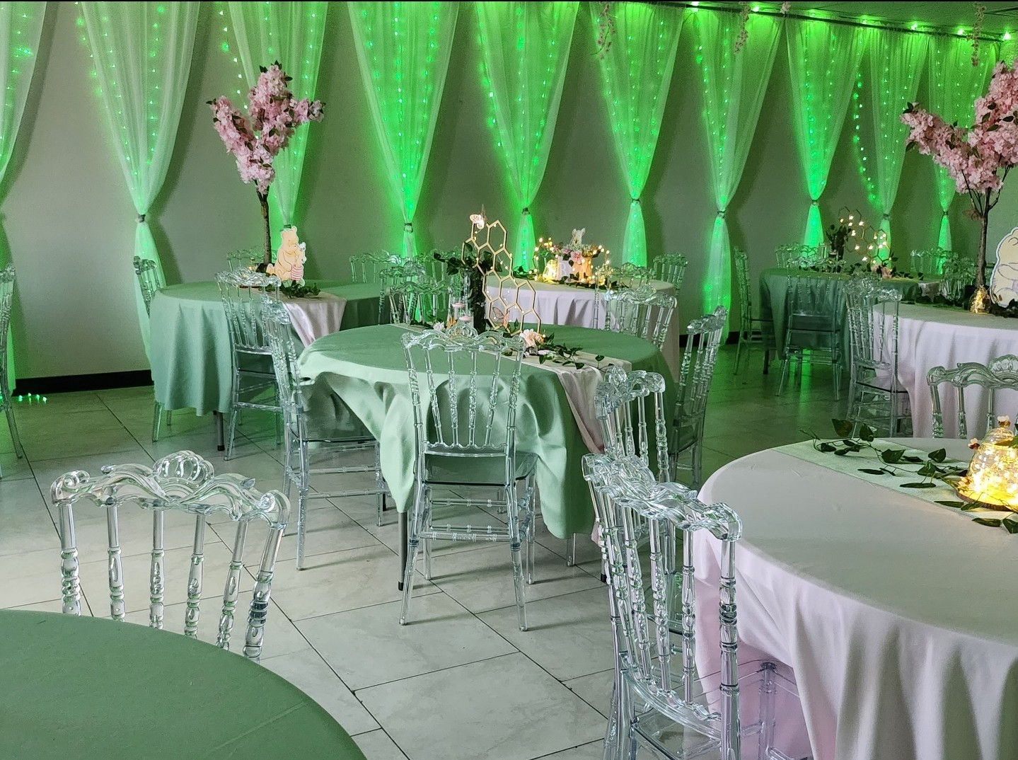 Elegant event space with green lighting, decorated tables with flowers, and transparent chairs, set for a celebration.