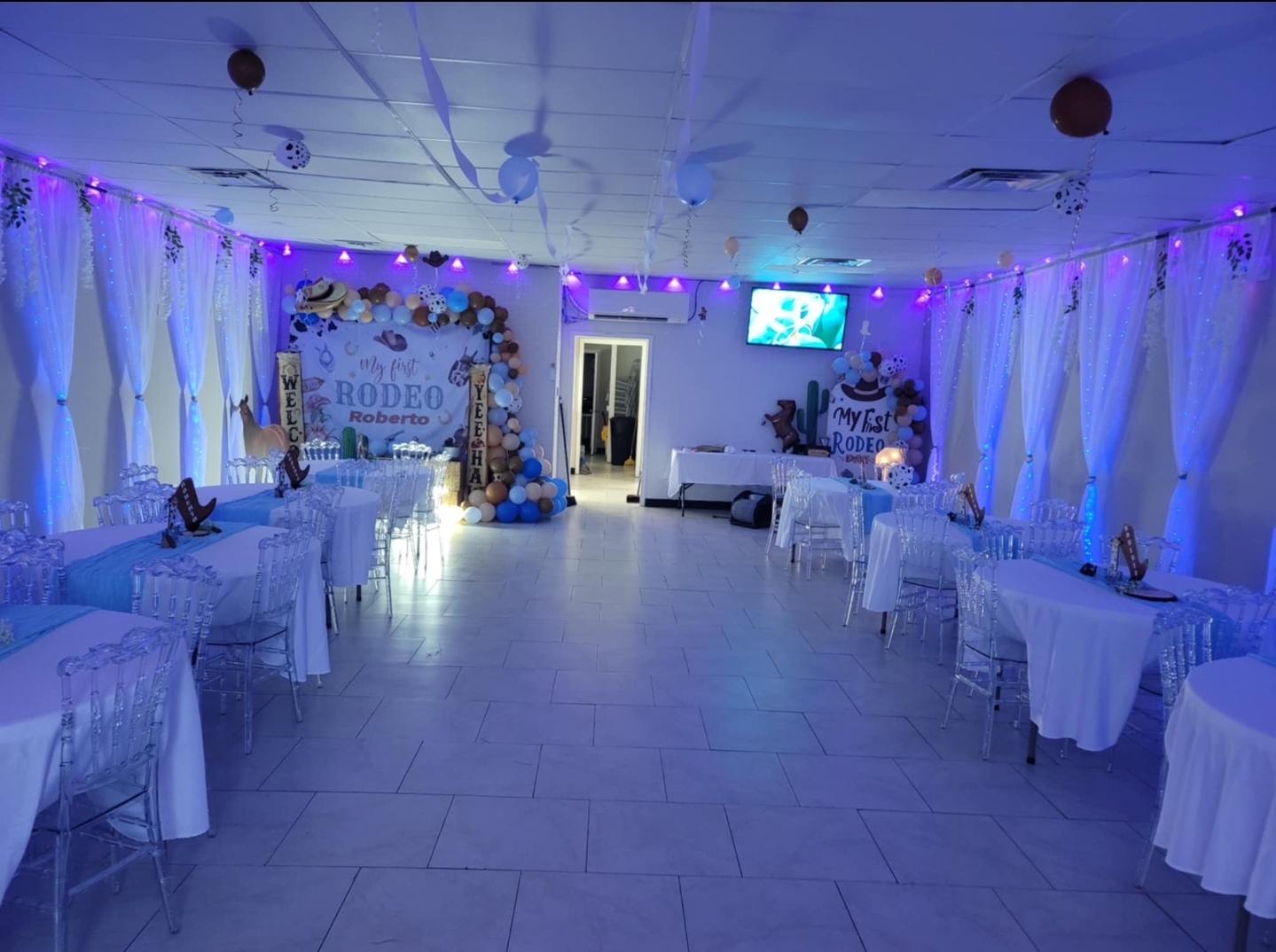 Event hall decorated with white curtains, purple string lights, tables with white tablecloths, and a decorated backdrop.
