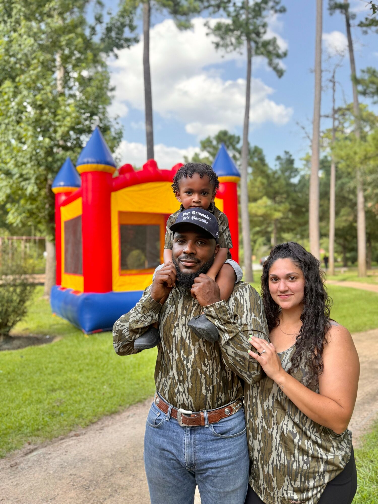 A man carrying a child on his shoulders and a woman standing beside them outdoors with a bounce house in the background.