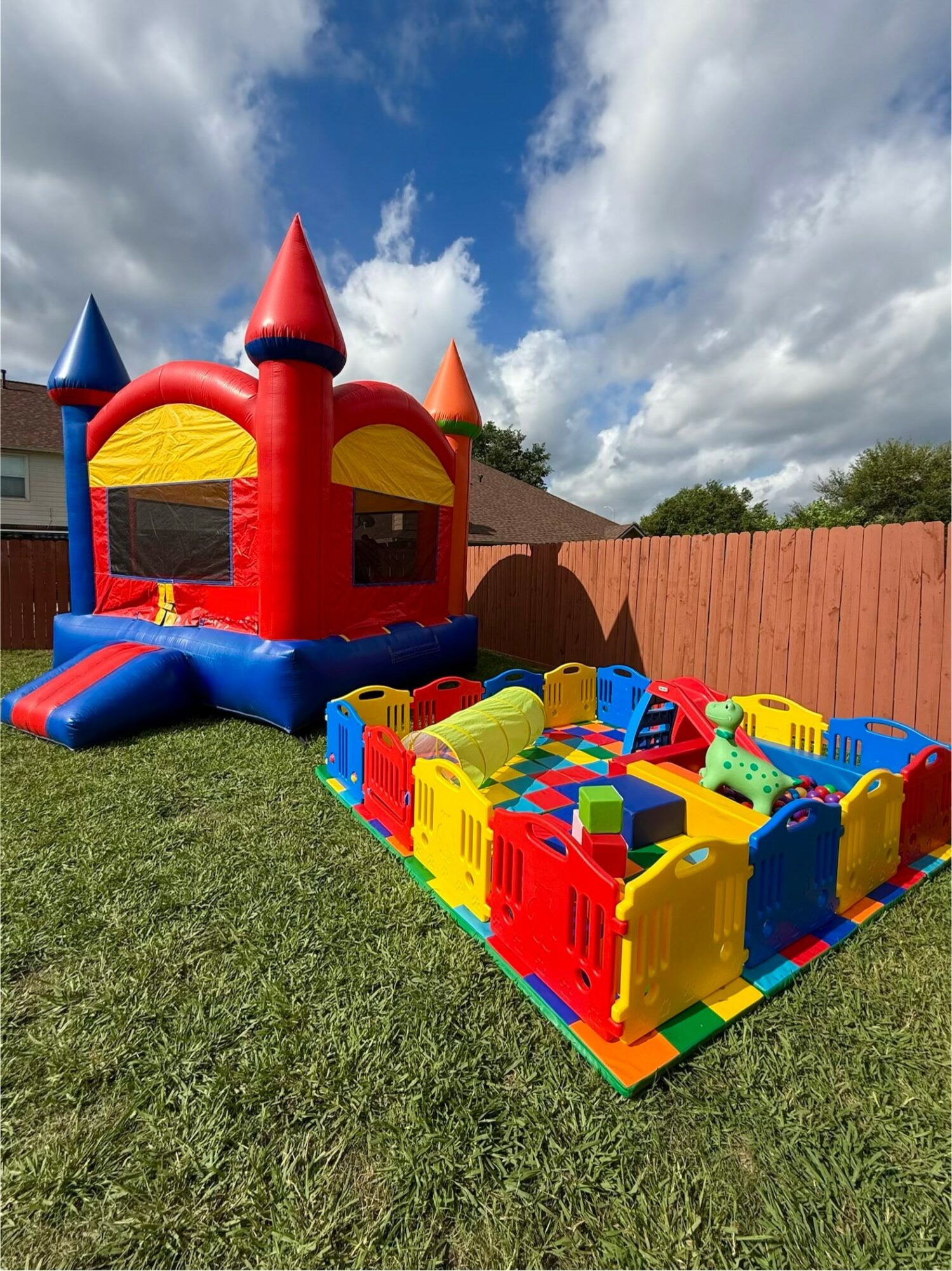 Colorful inflatable castle with turrets and a bounce area, set on a grassy yard with a wooden fence and blue sky.