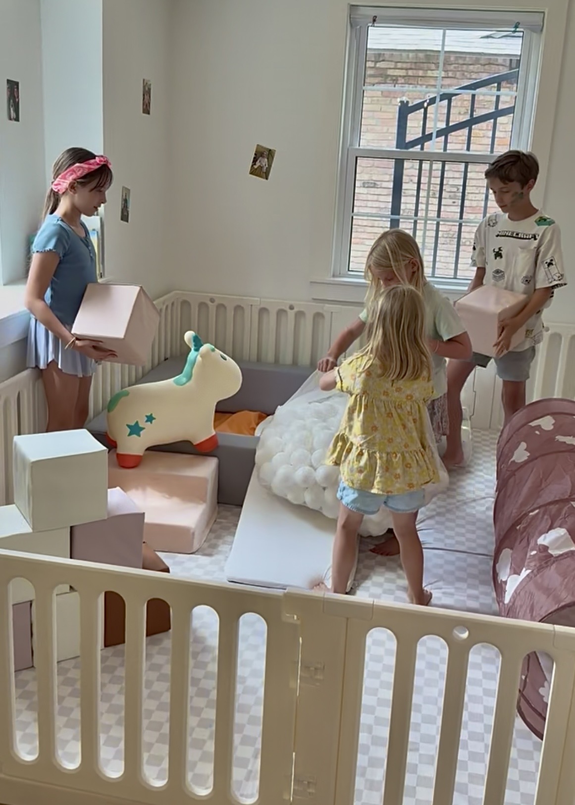 Three children playing in a room with a unicorn-shaped toy, boxes, and a window with bars, during daytime.