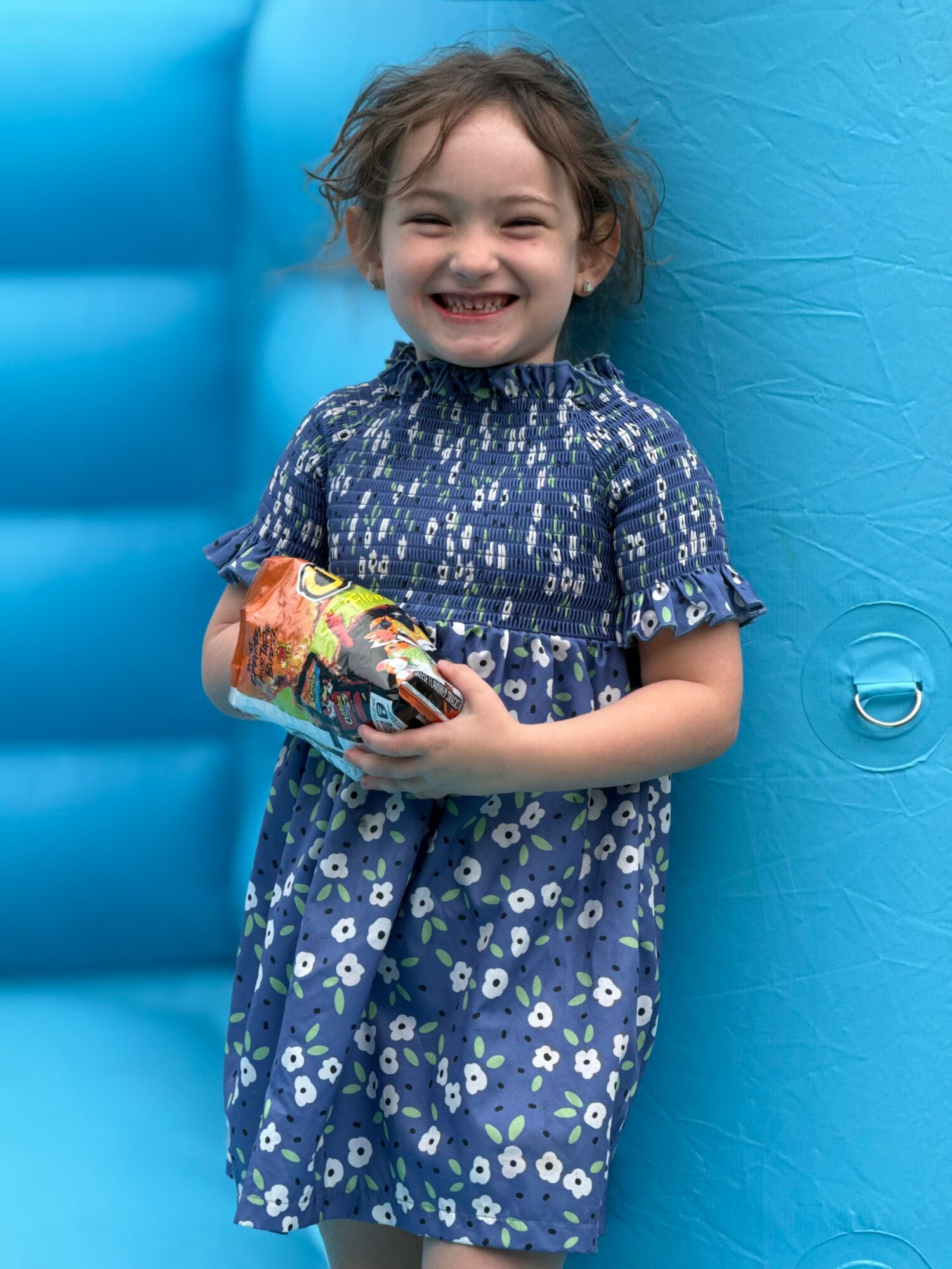 Smiling girl in a blue floral dress holding a toy, standing against a blue inflatable background.