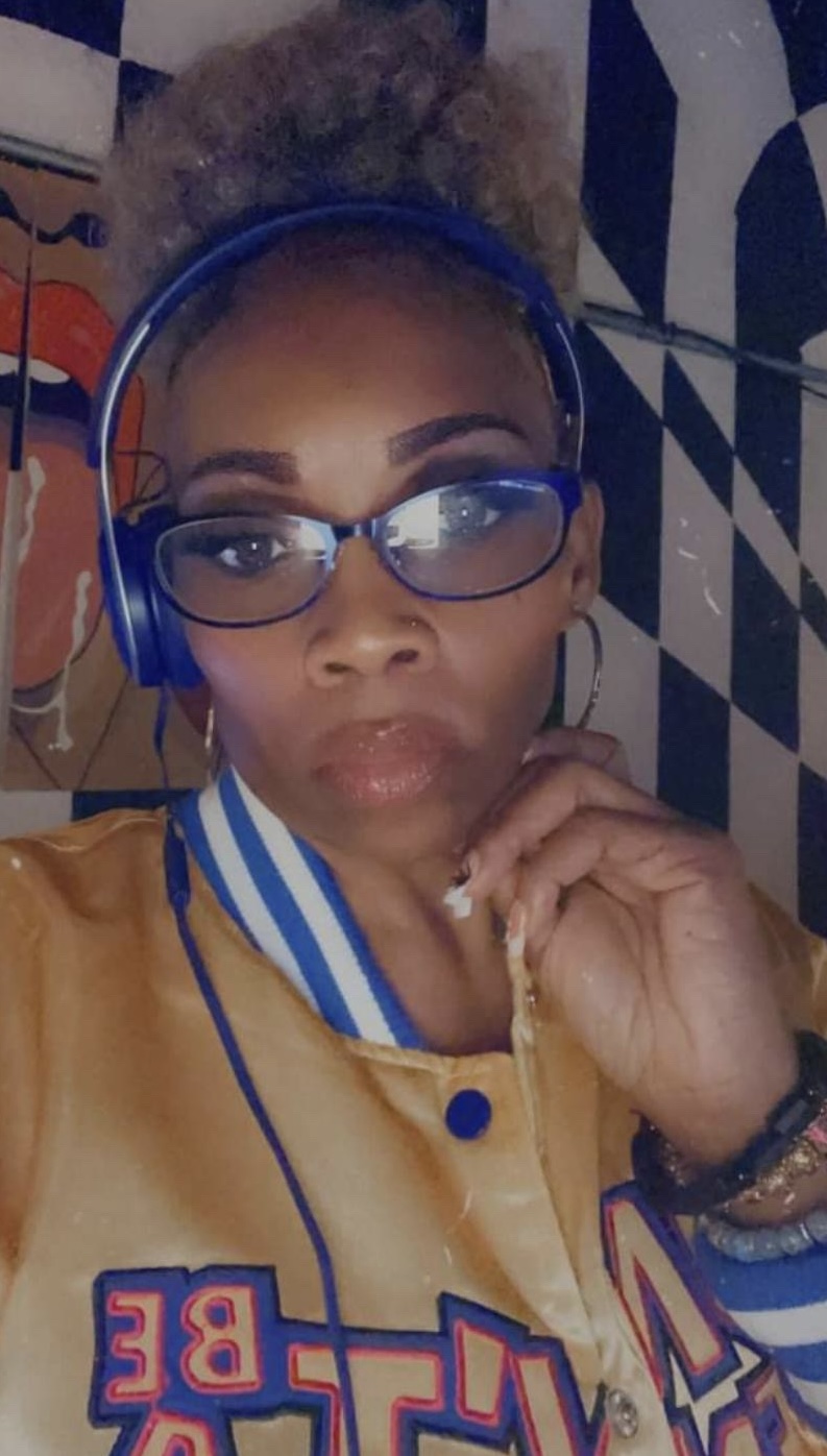 Woman with glasses and headphones, wearing a yellow jacket, resting her chin on her hand, in front of a patterned background.