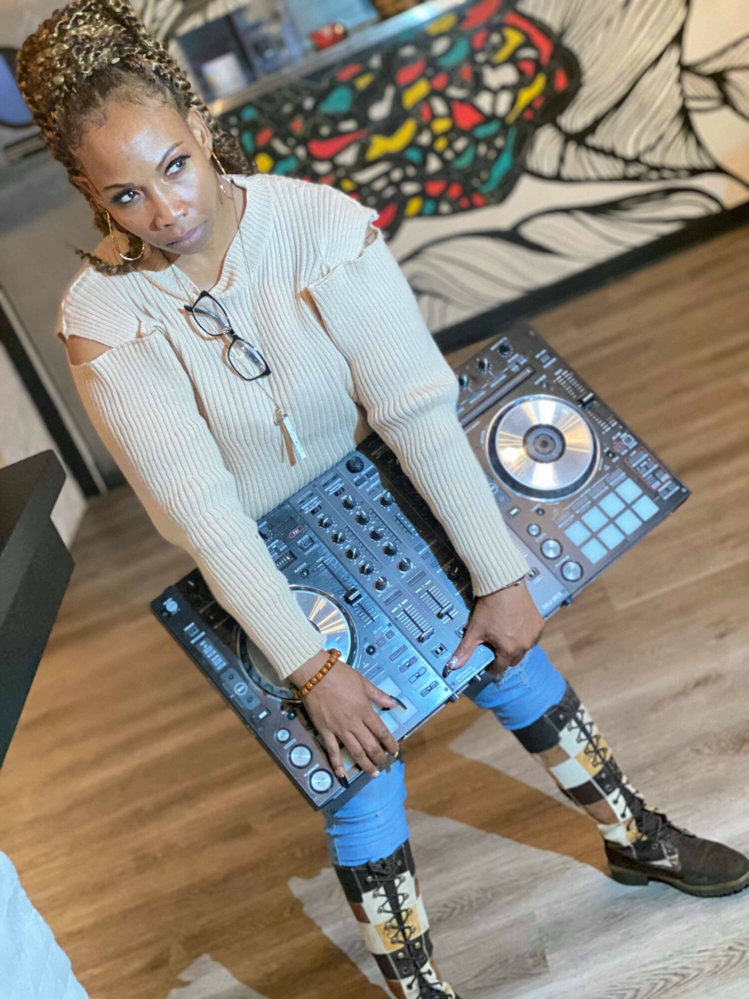 Woman standing with DJ equipment, looking at camera, in a room with patterned couch and wooden floor.