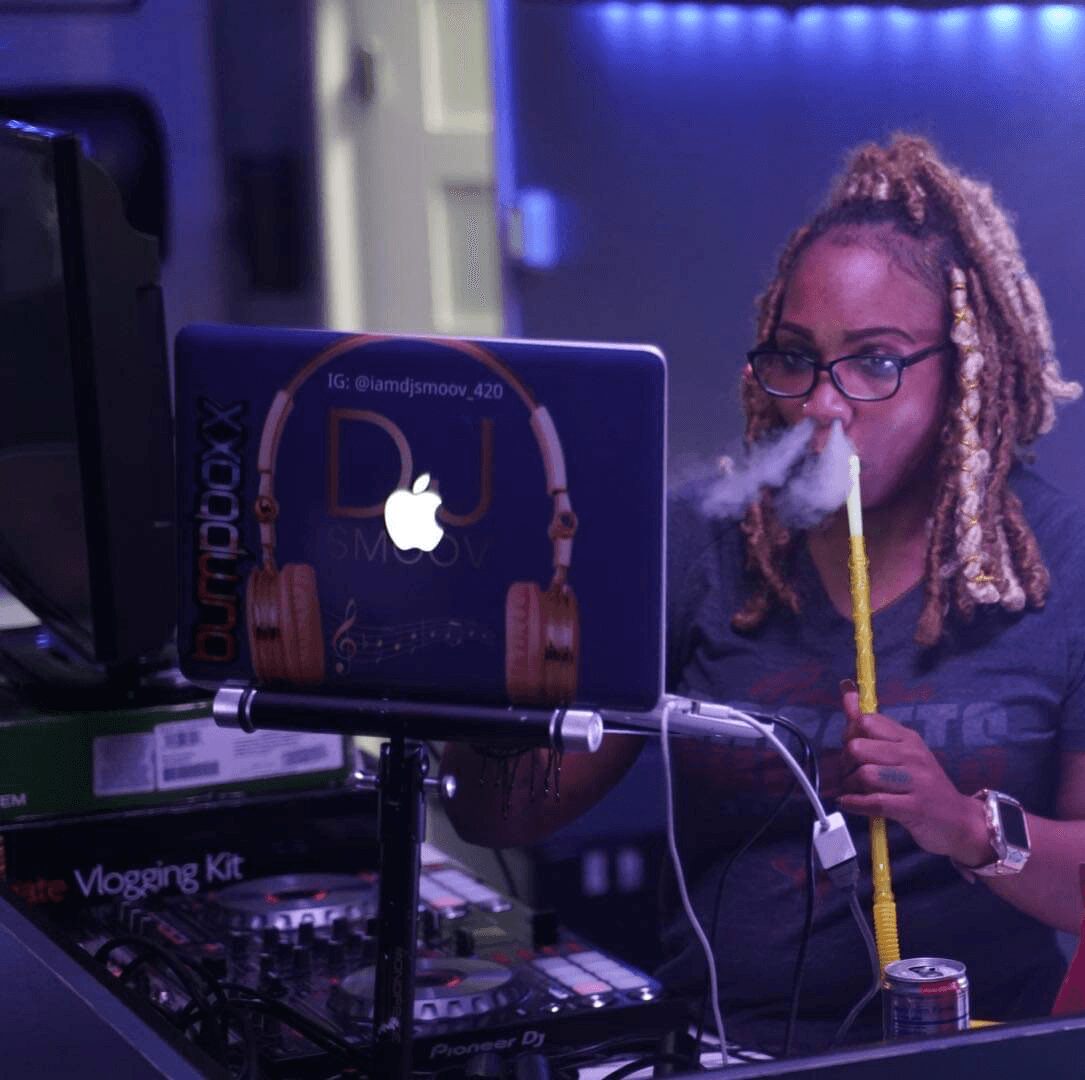 Woman with glasses and dreadlocks smokes a vape in a DJ setup, with a laptop and equipment, in a dimly lit room.