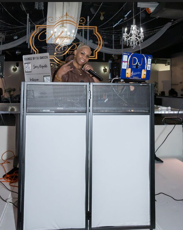 Person standing behind DJ booth with a microphone, smiling, in a decorated event space with chandeliers and a DJ screen.