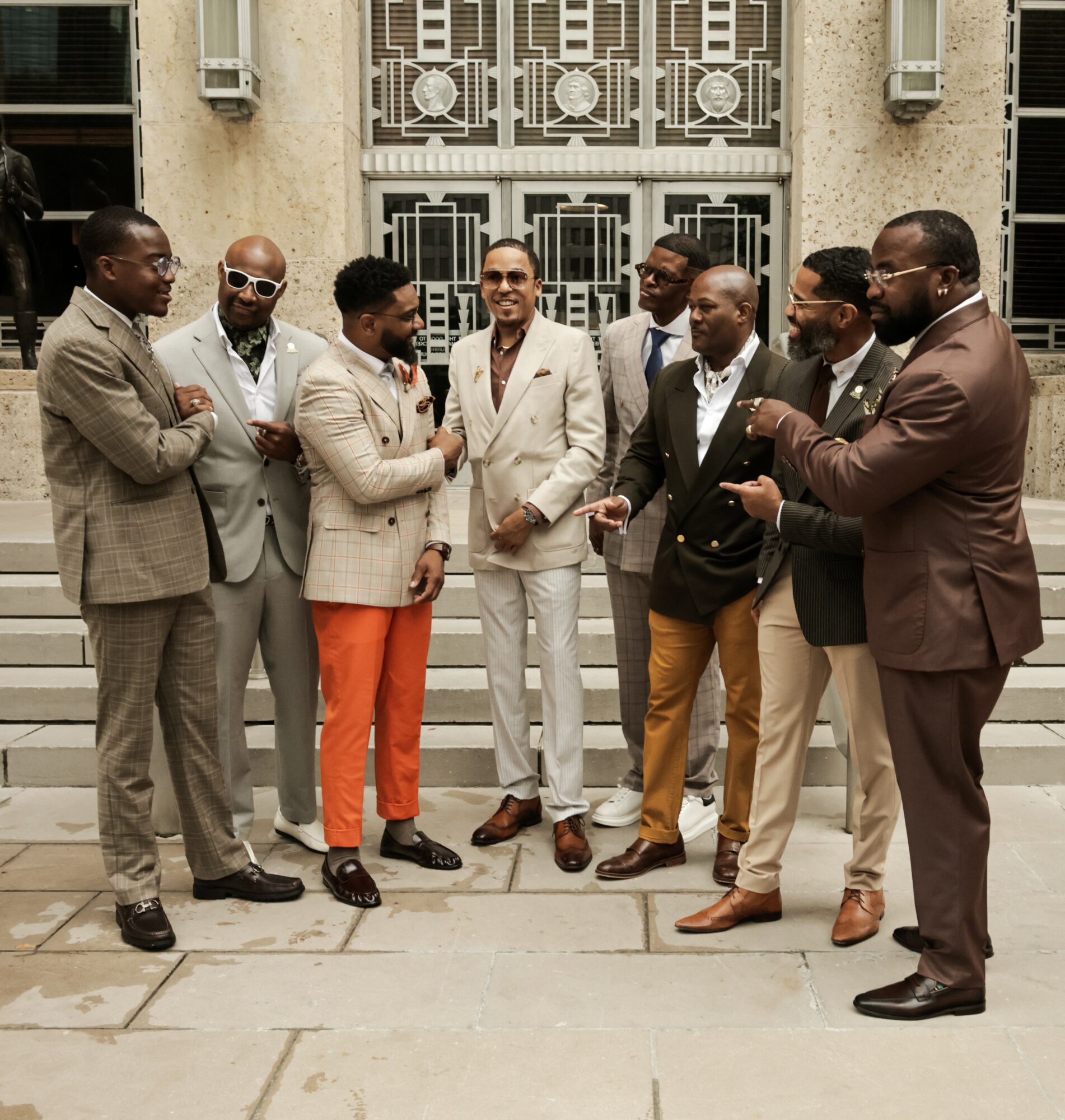 Group of eight men in suits standing outdoors, engaged in conversation, in front of a building with decorative metal doors.