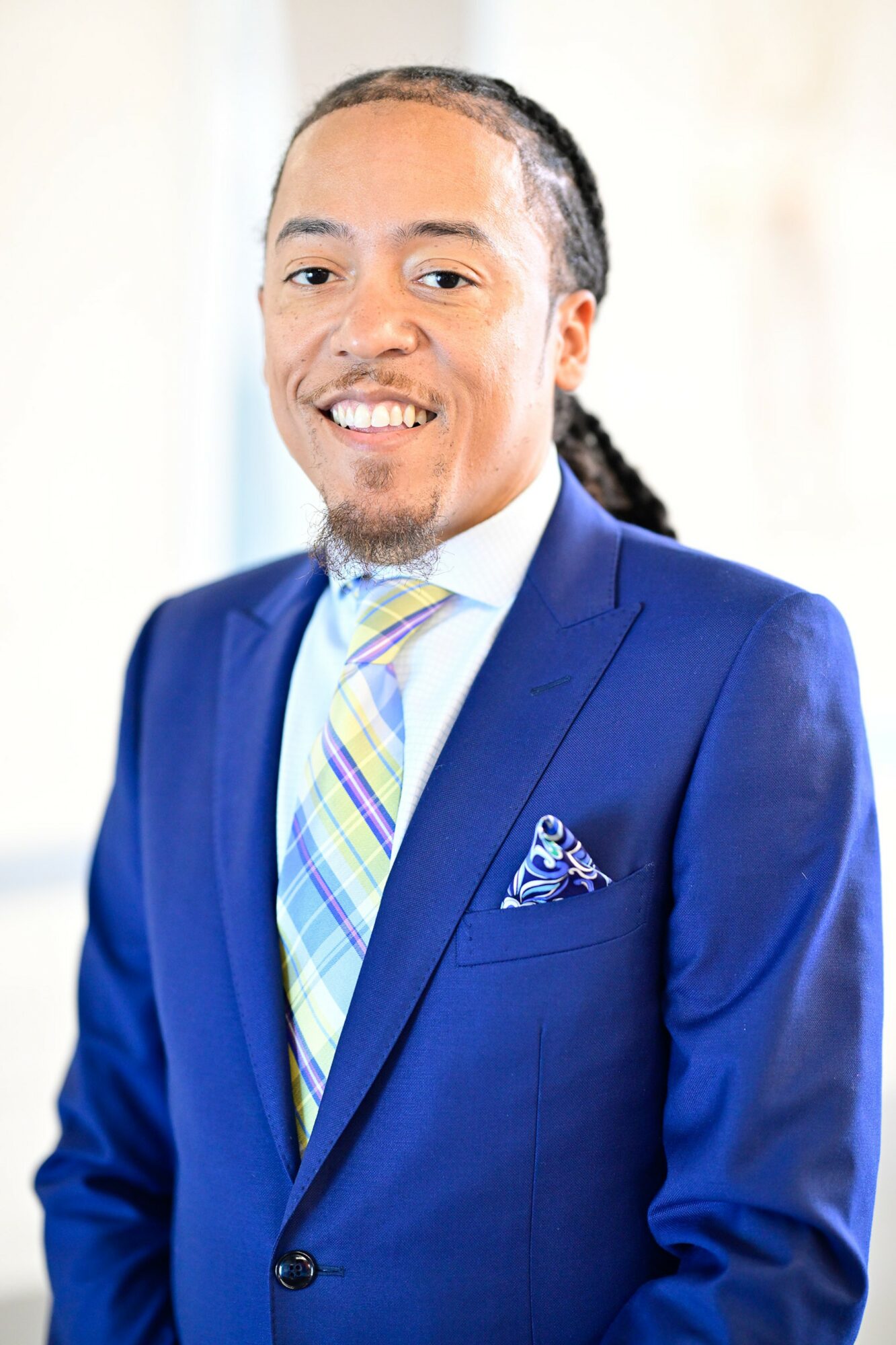 Man in blue suit with a colorful tie, smiling, with a pocket square, standing indoors with a blurred background.