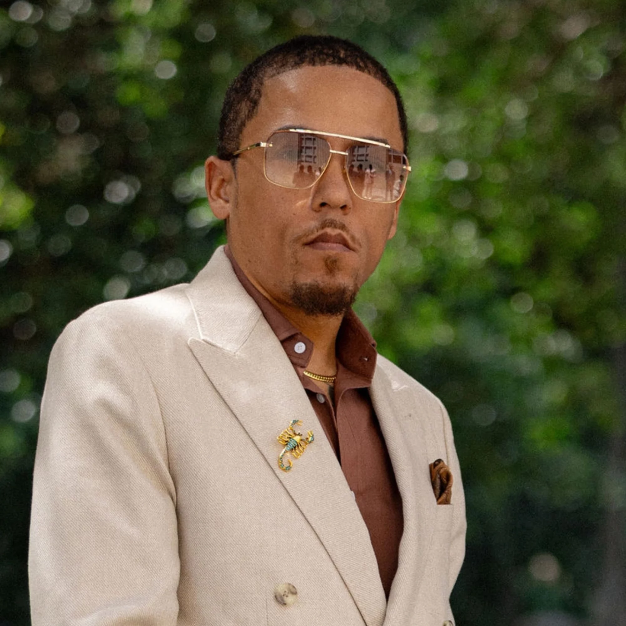 Man wearing sunglasses, beige blazer, and brown shirt outdoors with green foliage background.