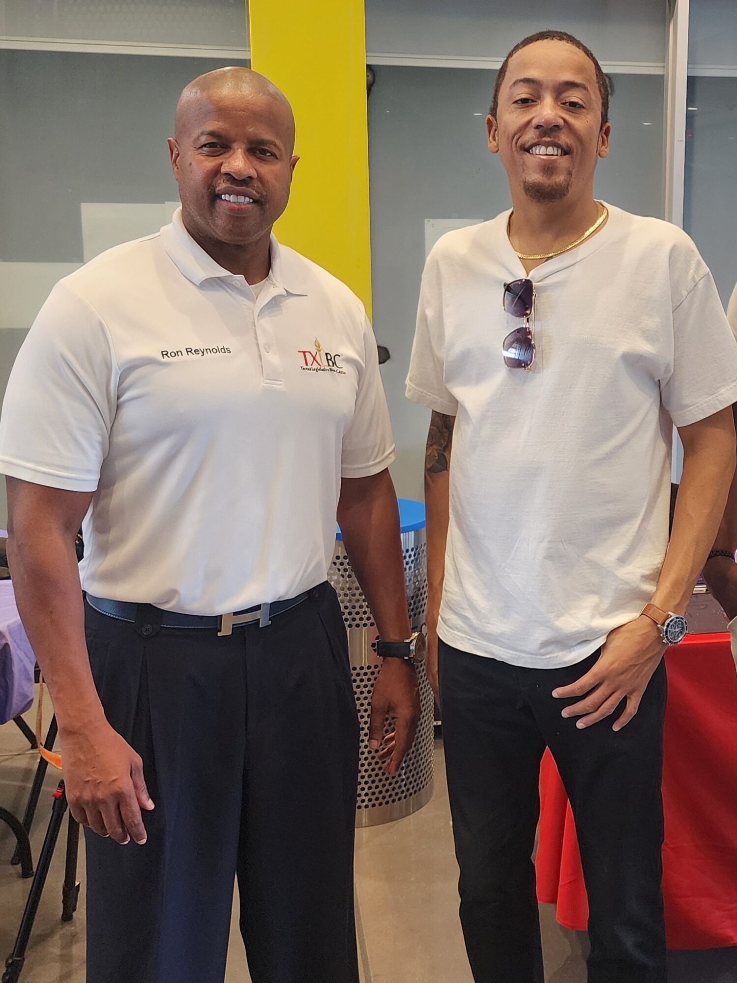 Two men standing indoors, smiling, one in a white polo shirt and the other in a white t-shirt, with a yellow wall behind them.