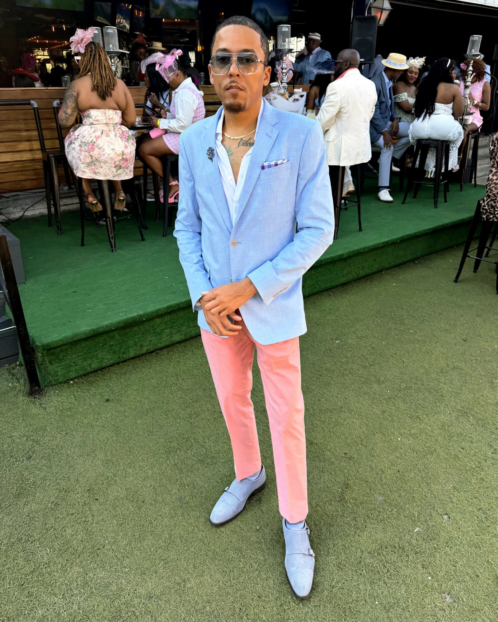 Man in light blue blazer, pink pants, and sunglasses standing on green surface at outdoor event with seated people in background.