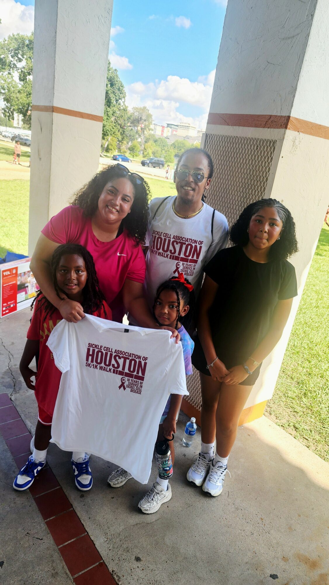 Four people, including children, stand outdoors near large white columns, holding a white Houston T-shirt, with a grassy area and trees in background.