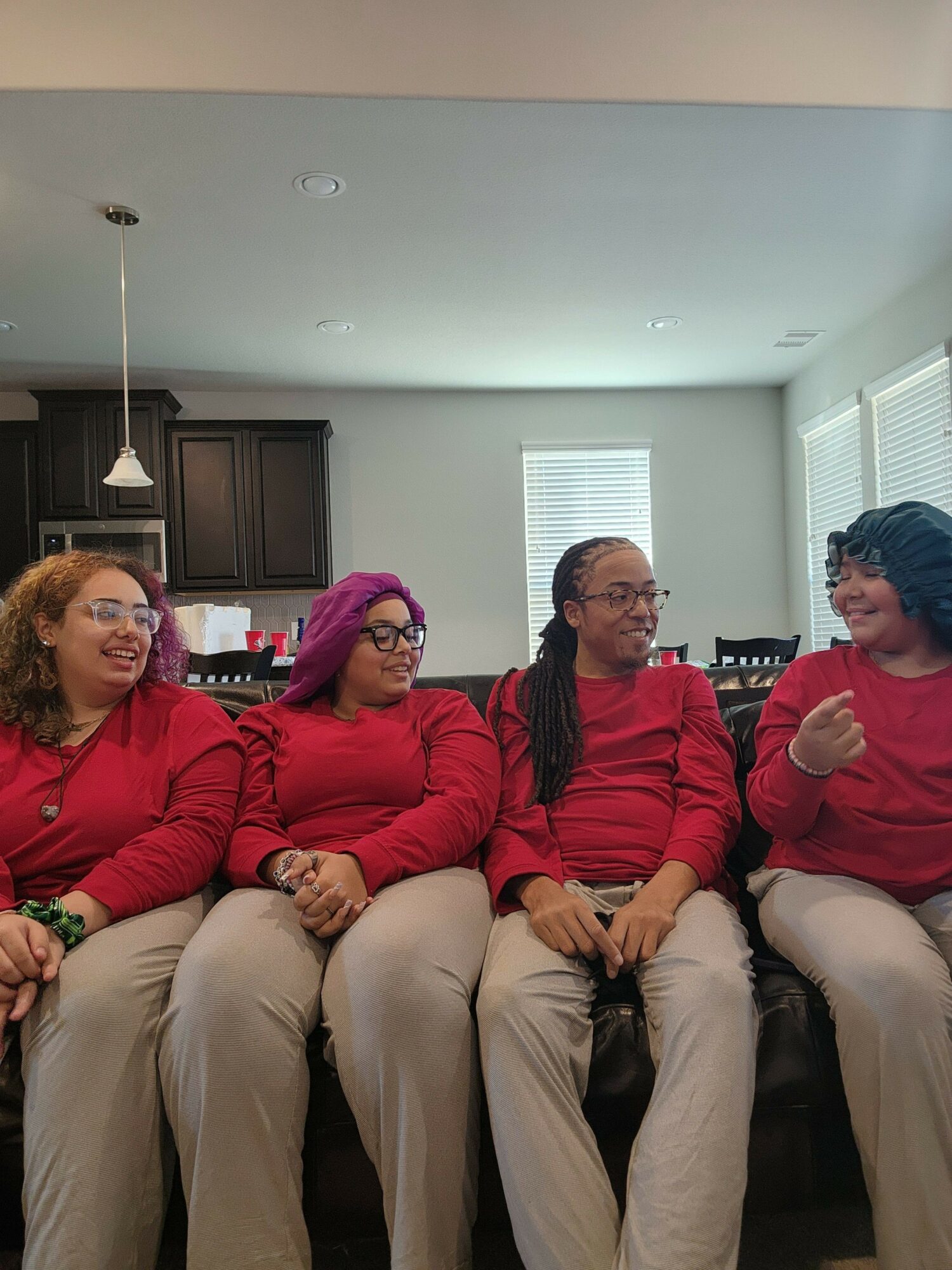 Four people sitting on a couch in a room with windows, two wearing red shirts, one with purple hair, and one with glasses.