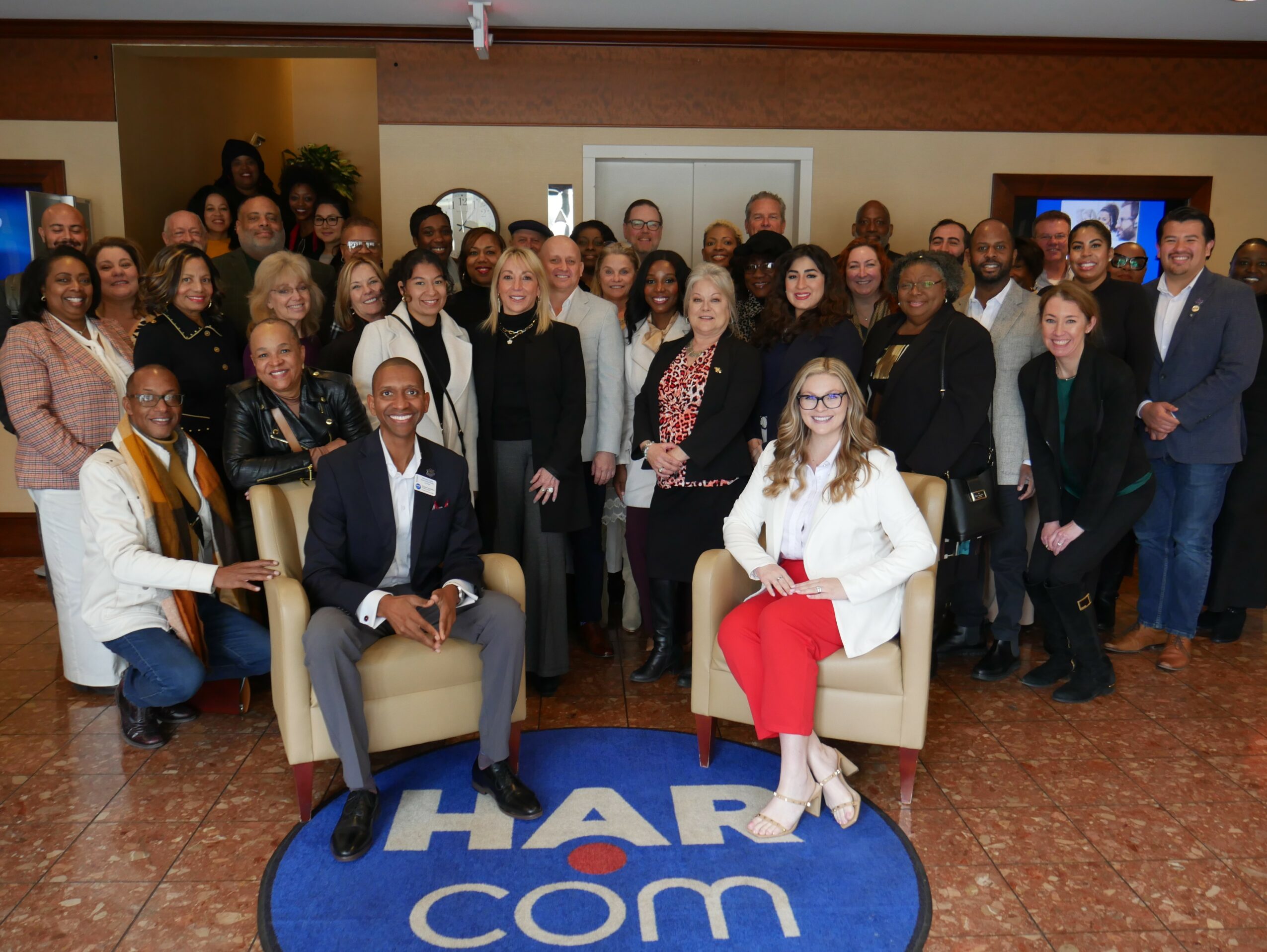 Group of diverse people gathered indoors, some seated and some standing, with a blue and white logo on the floor.