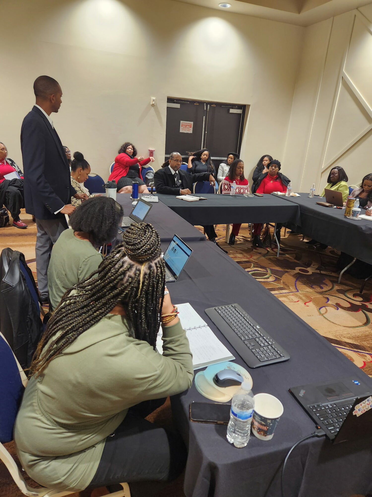 People seated around a U-shaped table in a conference room, some using laptops, with a person standing at the side.