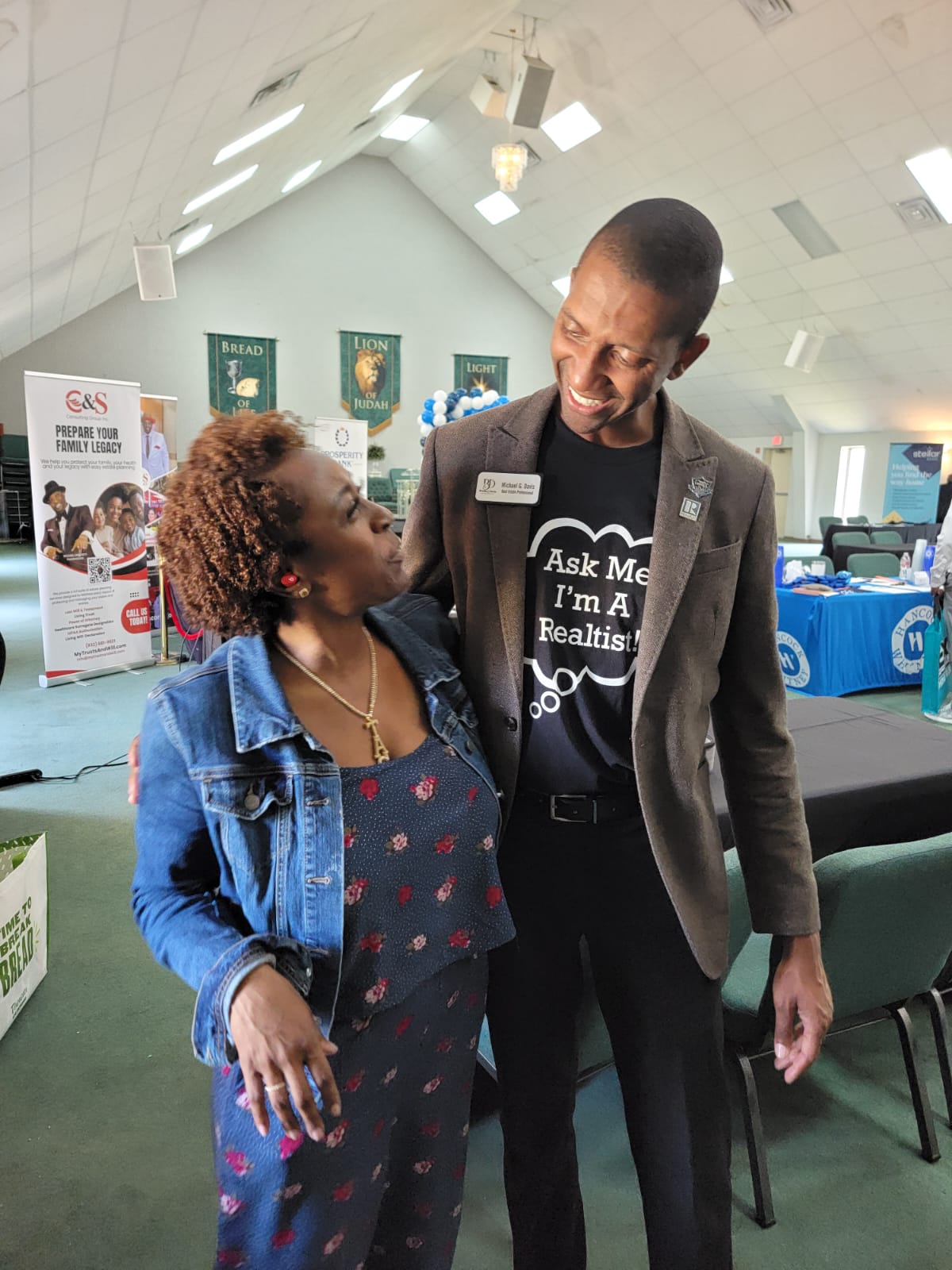 Two people talking indoors, one wearing a denim jacket and the other in a black shirt with white text, smiling at each other.
