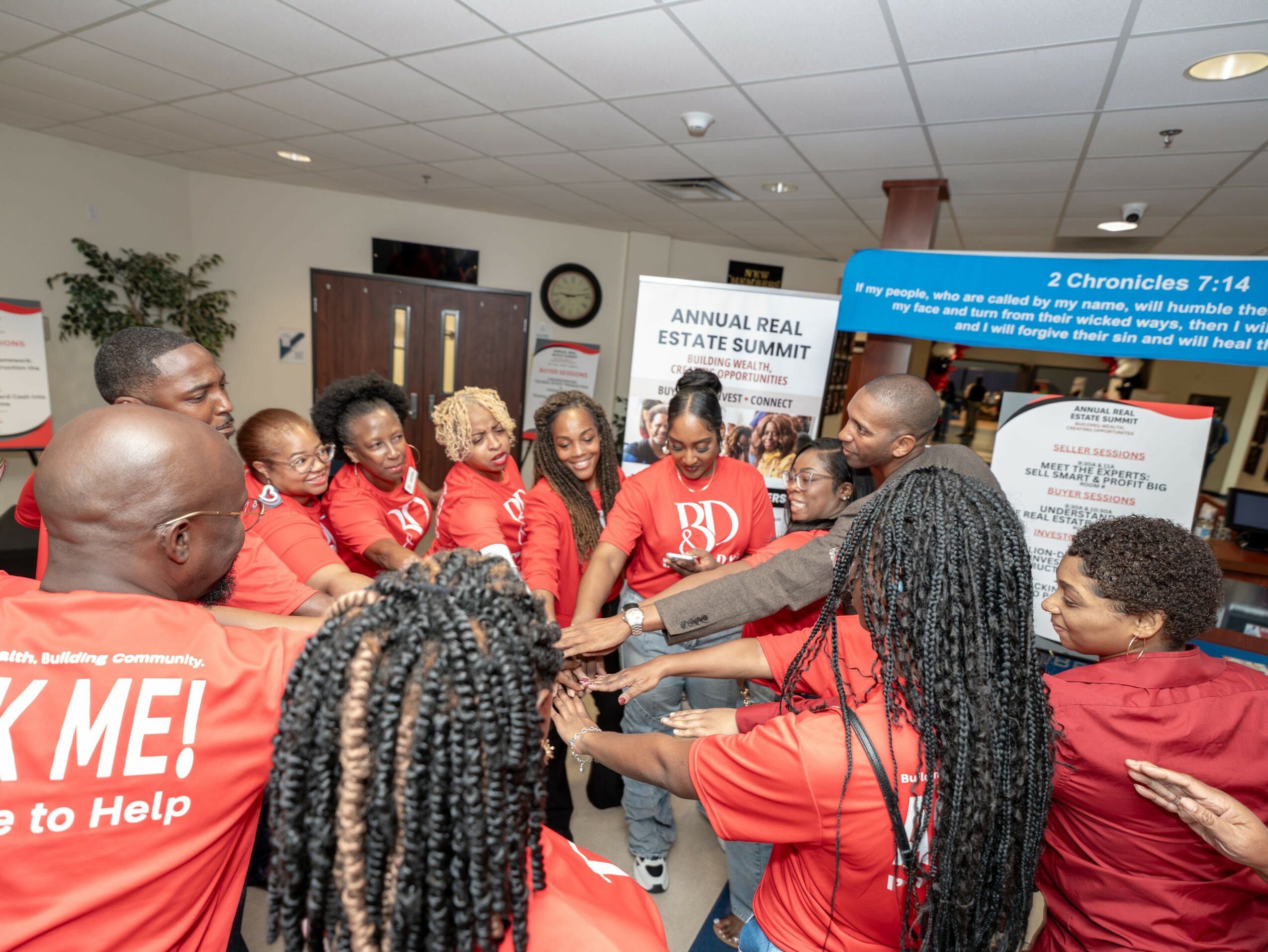 Group of people in red shirts gathered indoors, holding hands in a circle, participating in a team activity.