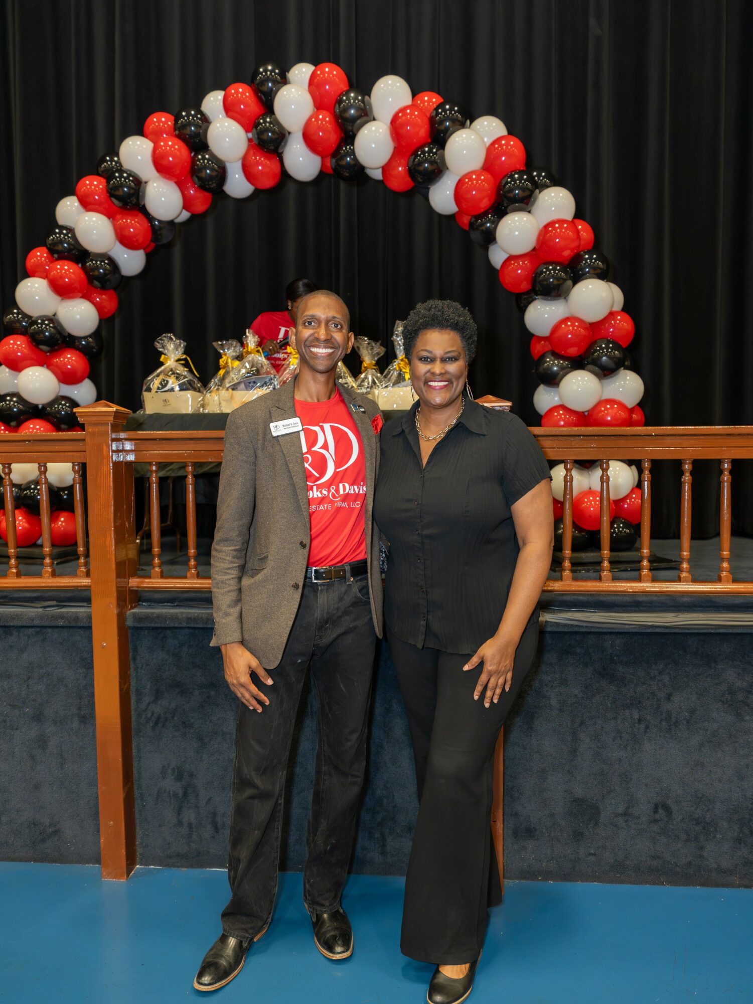 Two people standing in front of a balloon arch with red, black, and white balloons, smiling at the camera.