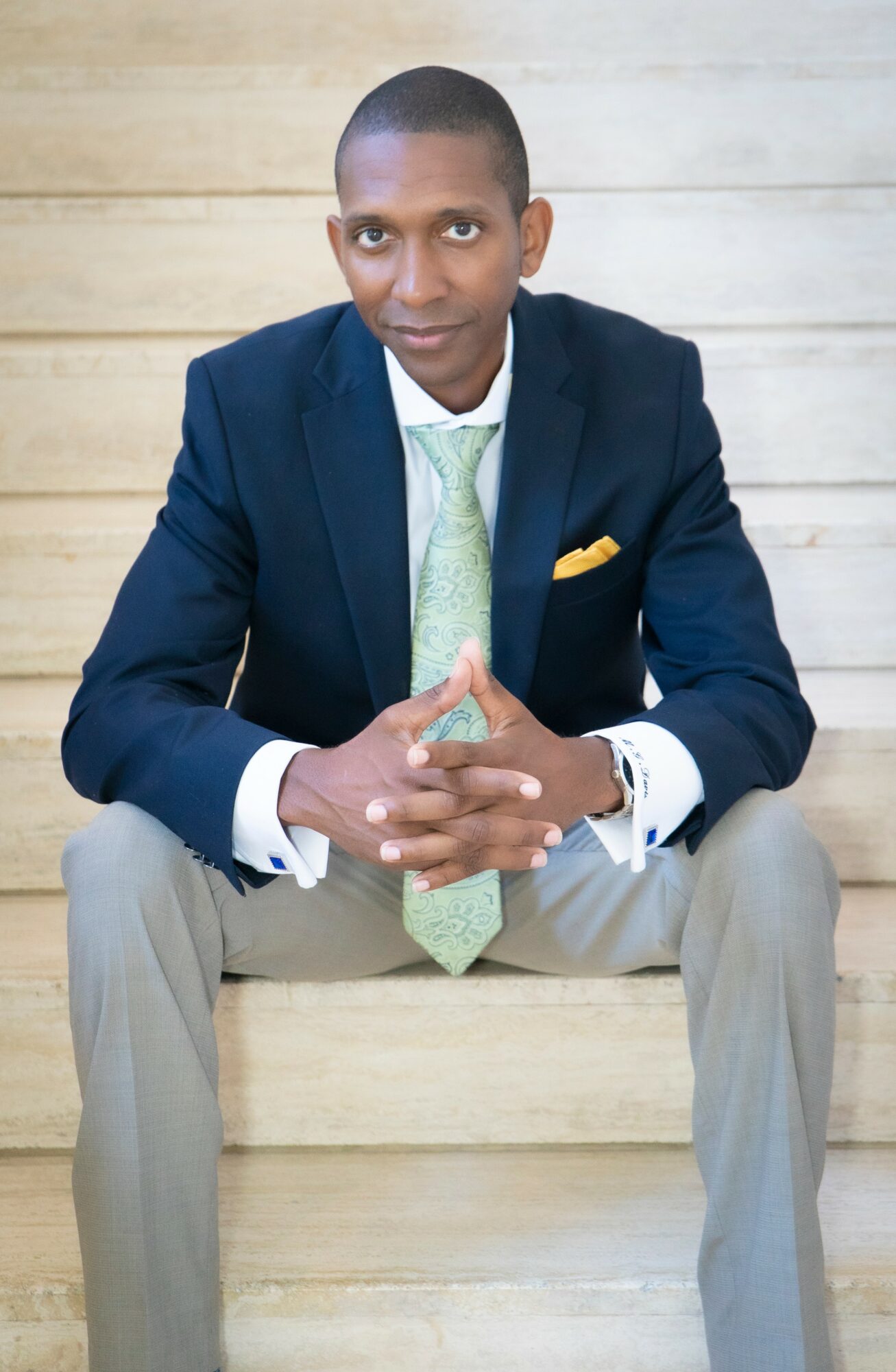 Man in a navy blazer, light green tie, and light gray pants sitting on a wooden bench with hands clasped.