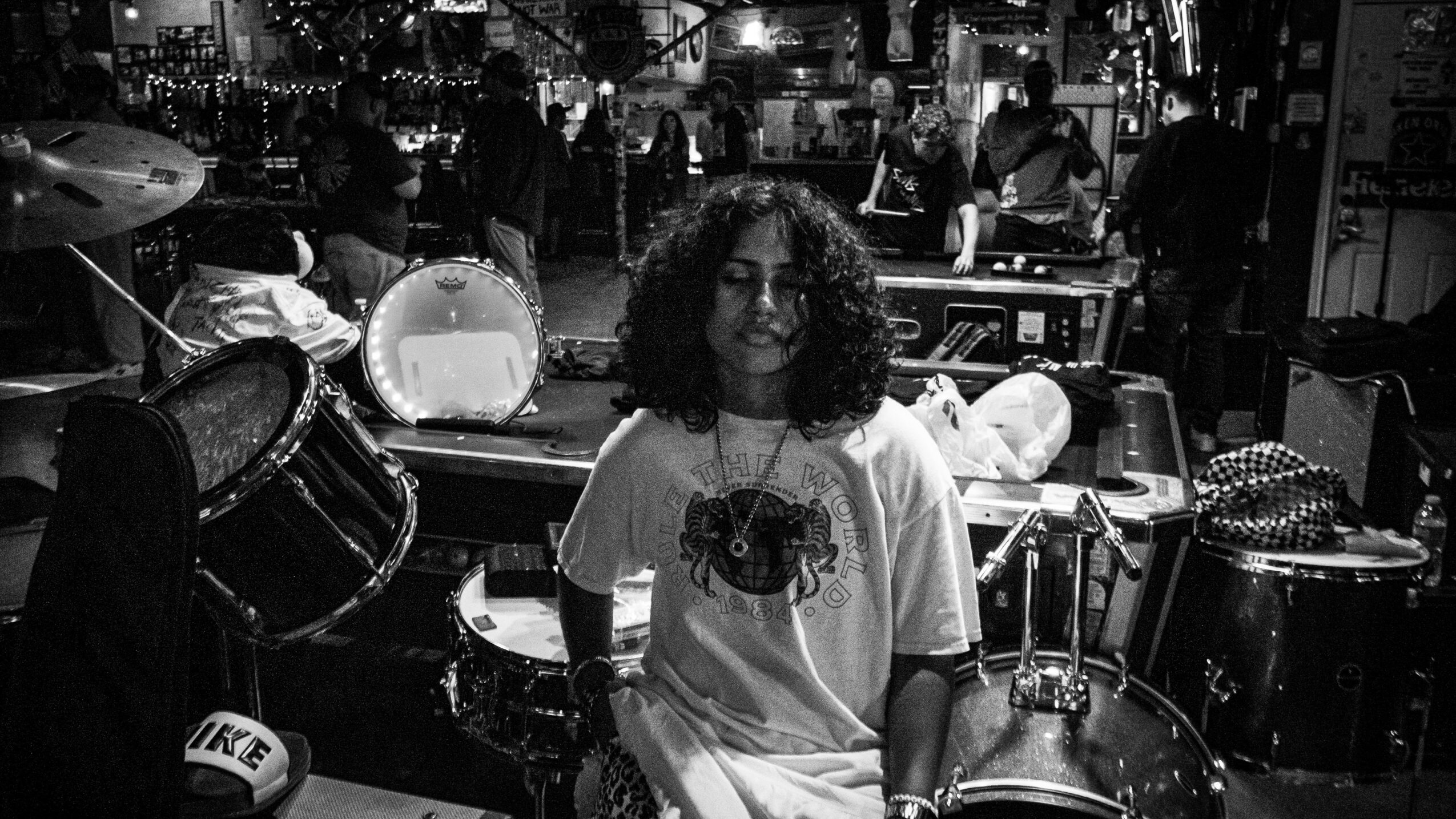 Person with curly hair sitting among drums and musical equipment in a busy indoor setting.