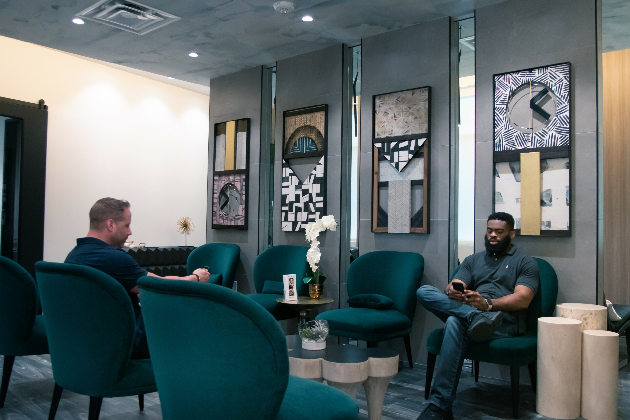 Two men sit in a modern lounge area with teal chairs and abstract wall art, one using a phone and the other looking away.