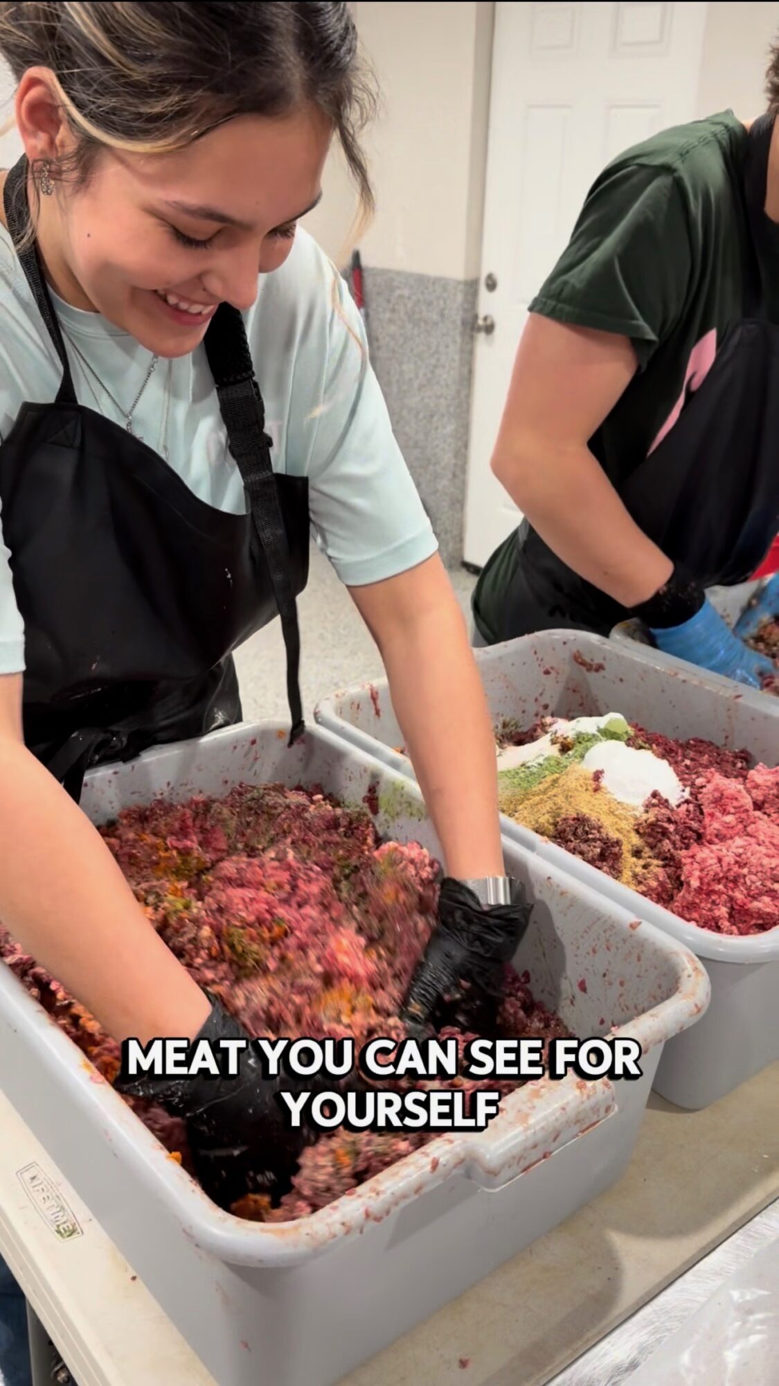 Two people preparing ground meat in large containers, smiling, in a kitchen setting.