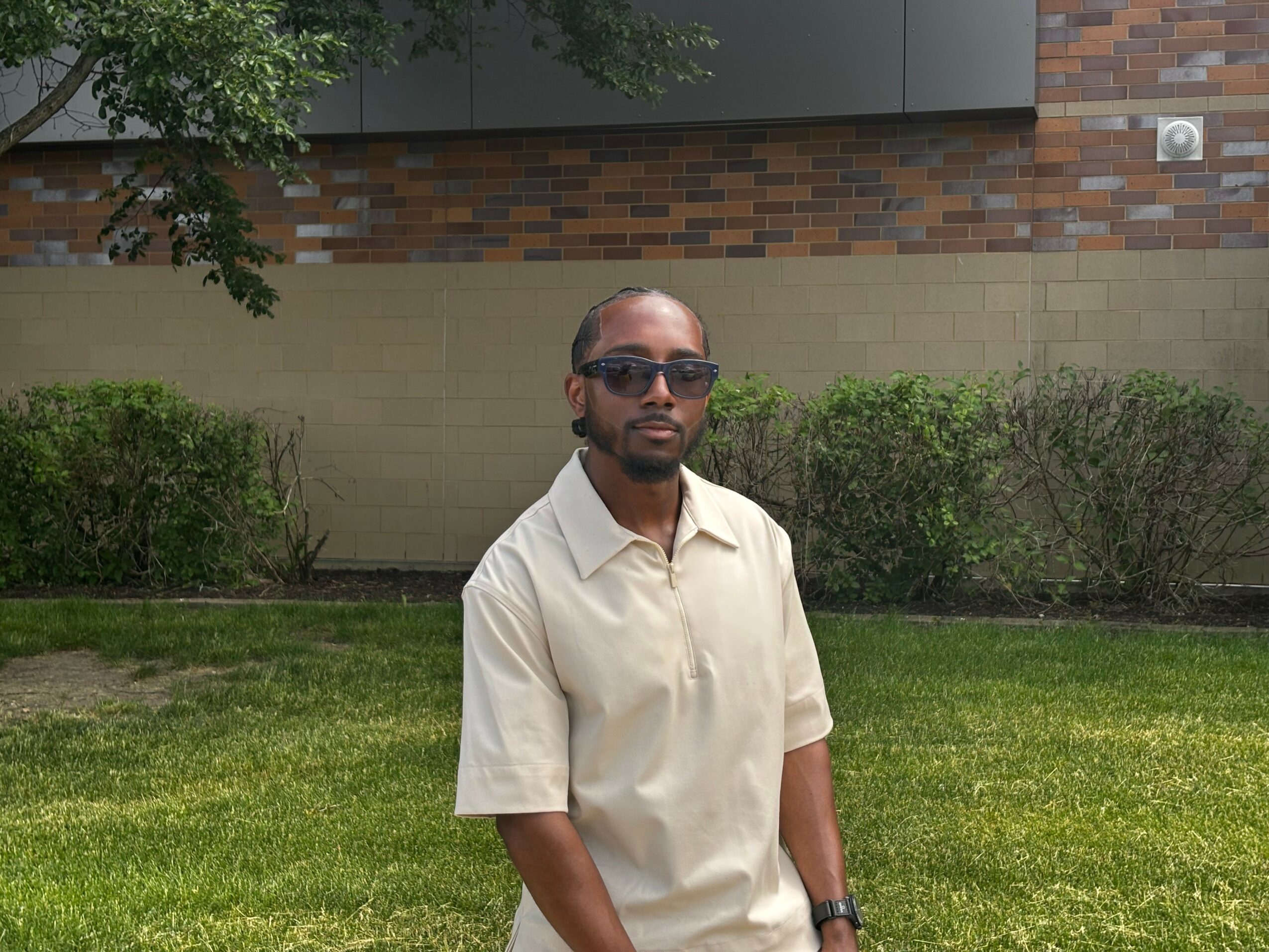 Man wearing sunglasses and a beige shirt standing outdoors with green bushes and a brick wall behind him.