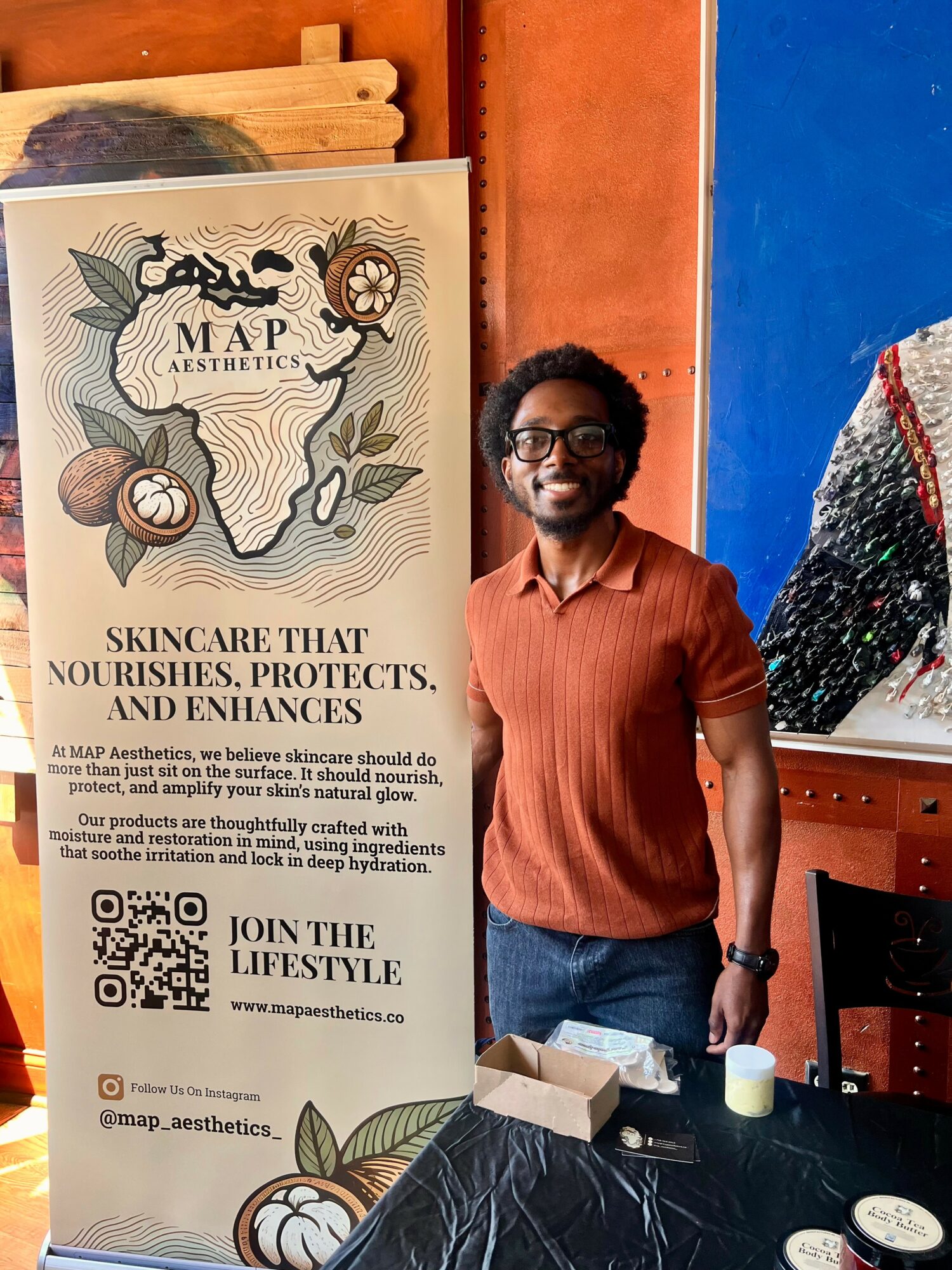 Smiling man with glasses and curly hair stands next to a vertical banner promoting skincare and aesthetics, in a room with colorful walls.