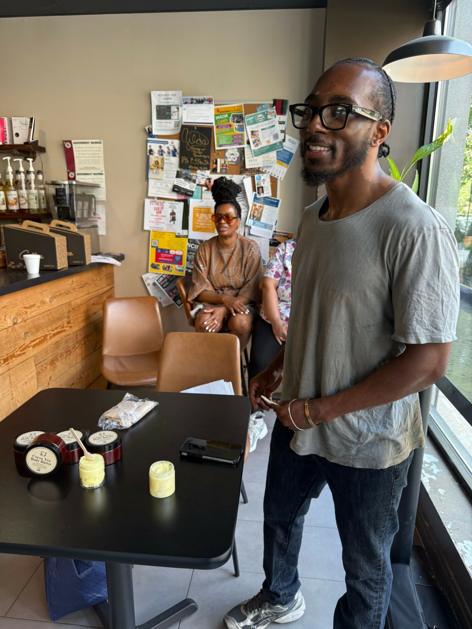 Two men standing in a room with a bulletin board, one wearing glasses and a gray shirt, the other in a brown shirt, with a table and various items.