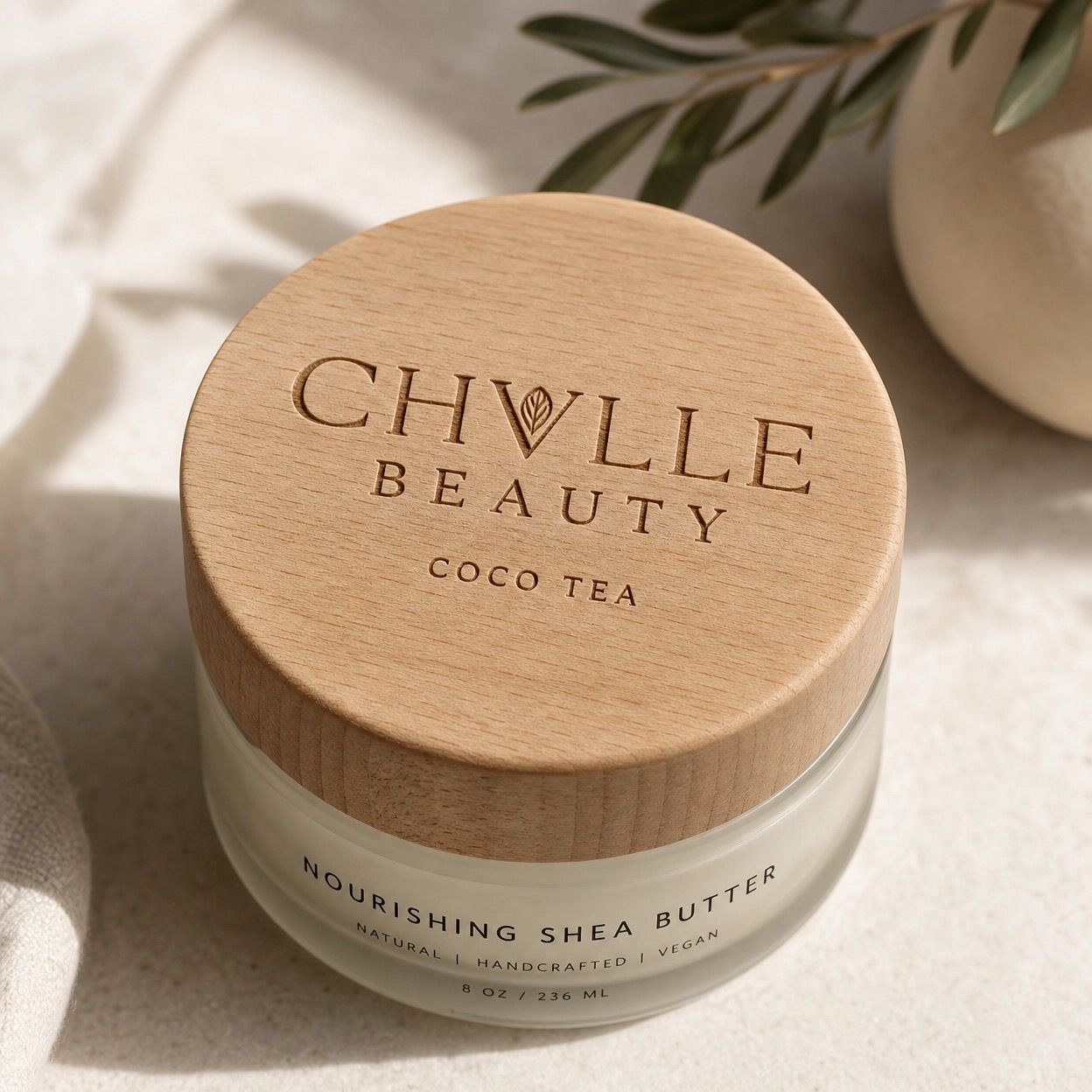 Round container with wooden lid labeled 'Chullie Beauty Coco Tea' and 'Nourishing Shea Butter' on a light surface with plant leaves nearby.