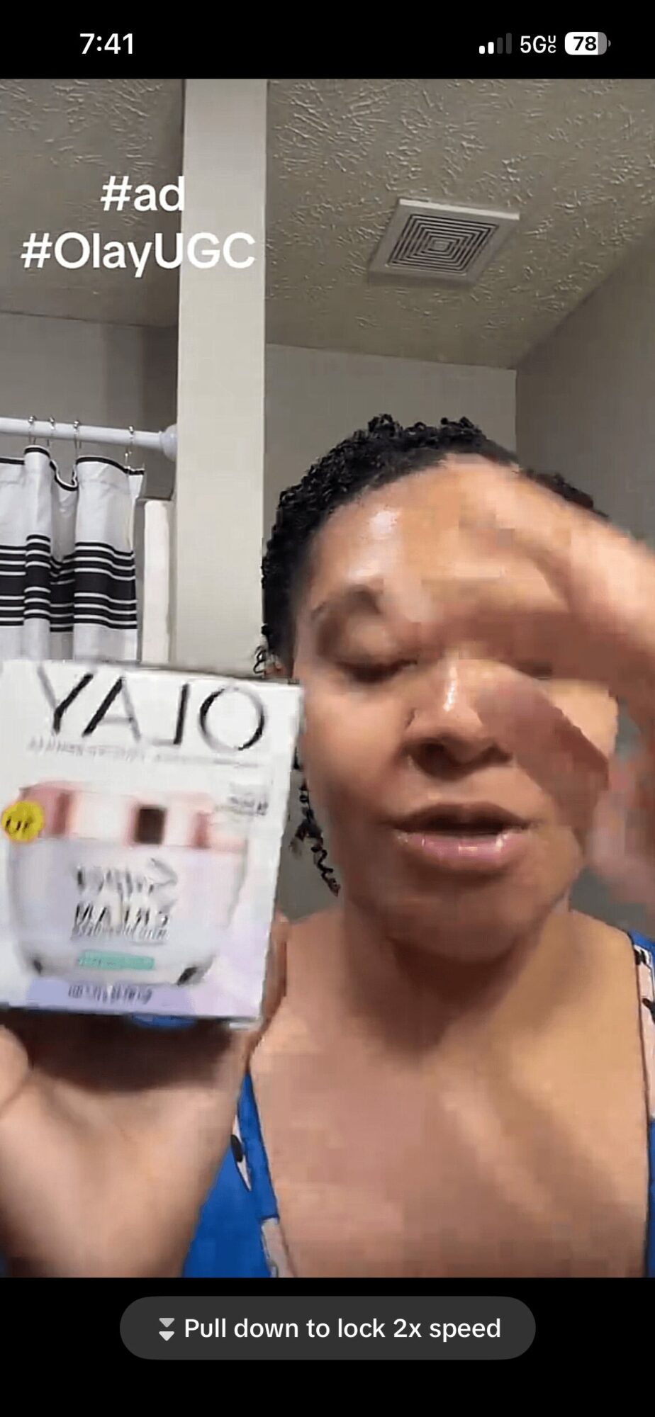Woman holding a product box, speaking, with a bathroom background, ceiling vent visible, wearing a blue top.