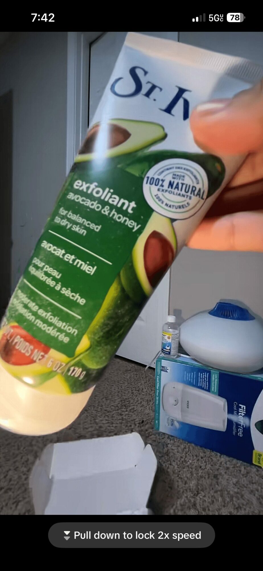 Hand holding a tube of exfoliant cream with a green and white label, in front of a window and a blue container.