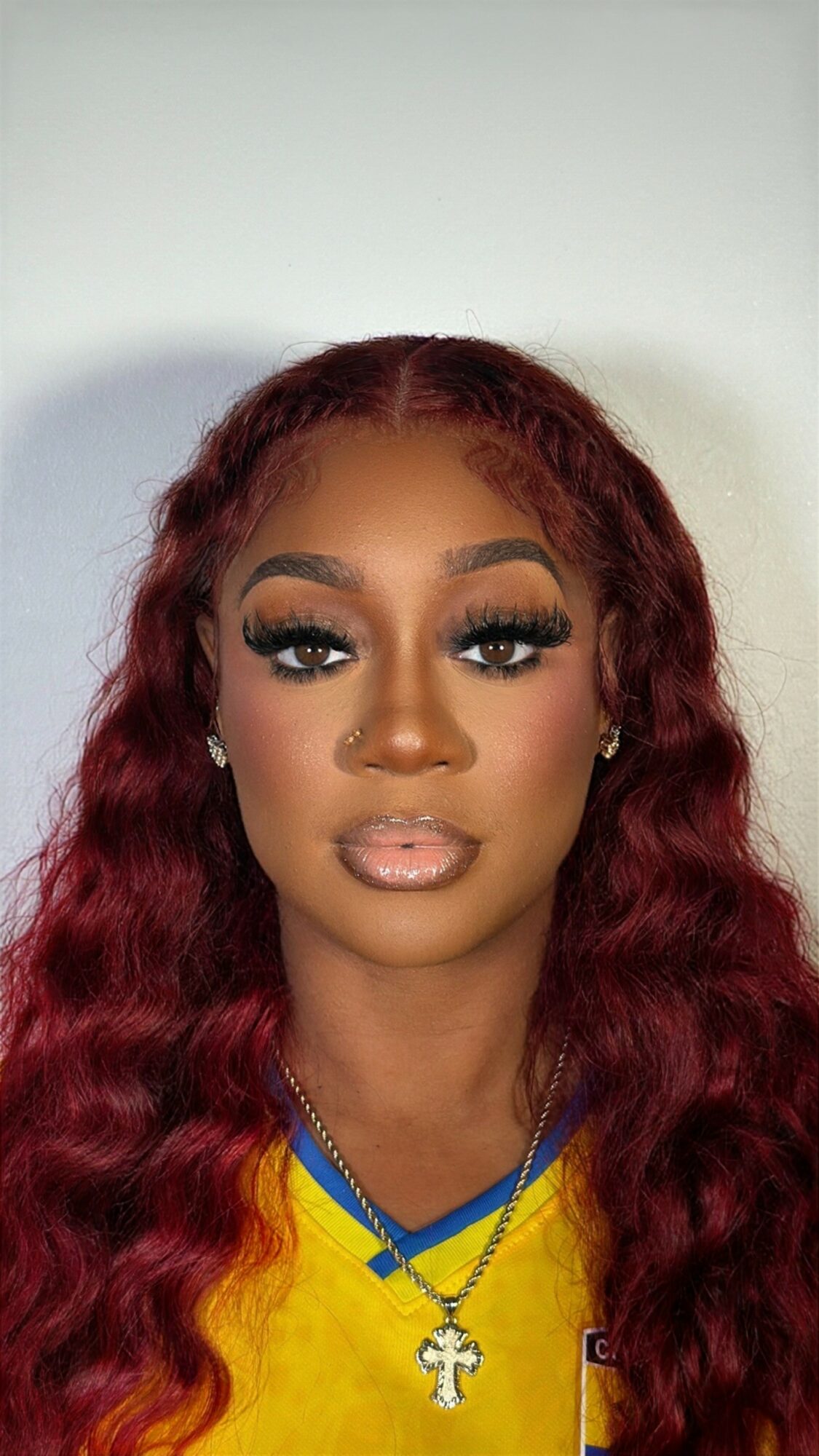 Portrait of a woman with long, wavy red hair, makeup, and a yellow top, against a plain background.