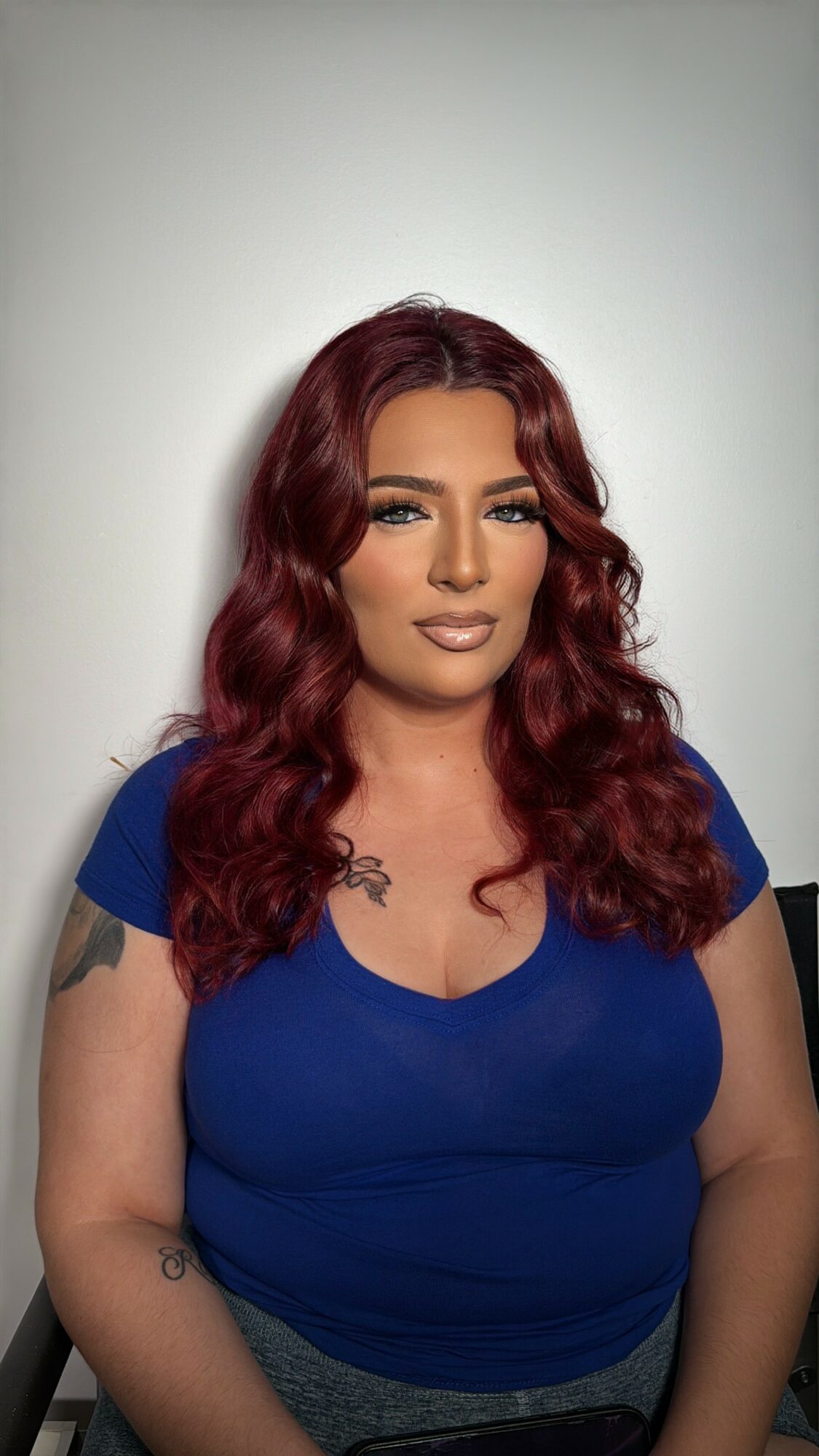 Woman with long wavy red hair wearing a blue top, standing against a plain light background.