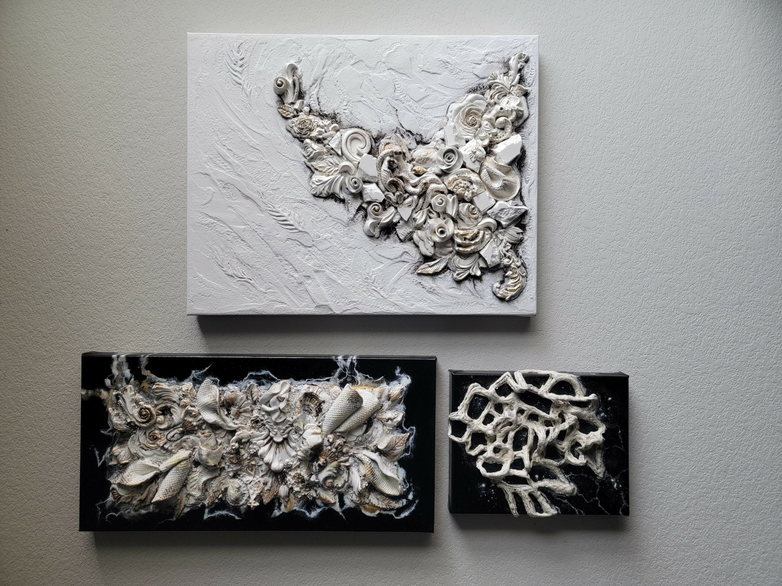 Three textured abstract art pieces on a wall, one above two smaller ones, with intricate patterns and a monochromatic color scheme.