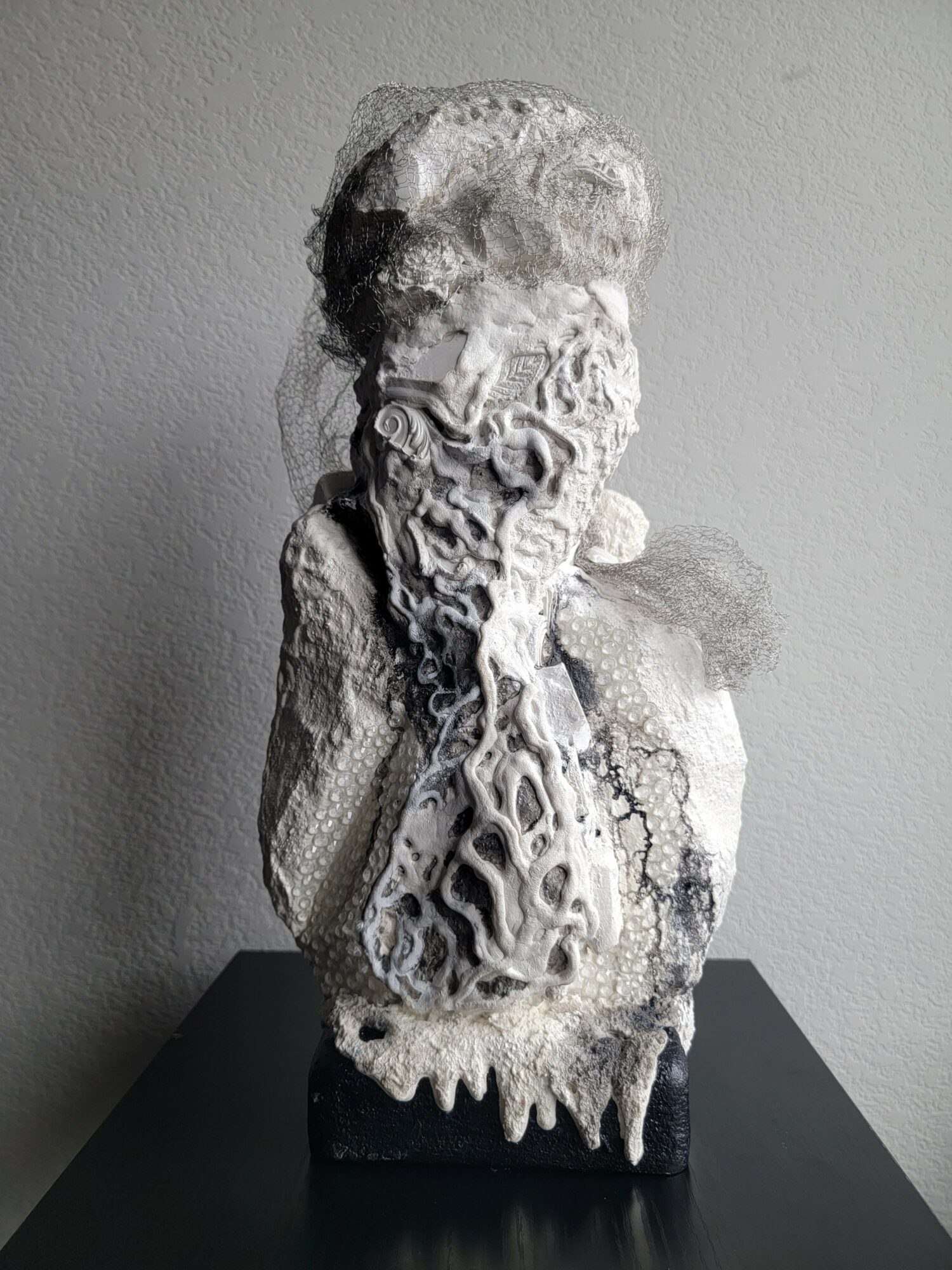 Sculpture with intricate, layered textures on a black base against a plain wall.